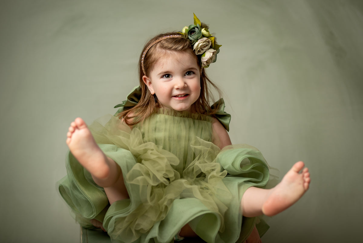 A young girl stands barefoot on a green box wearing a light olive green tulle dress with layered ruffles and oversized bow-tied shoulder straps, posing against a matching green backdrop.

