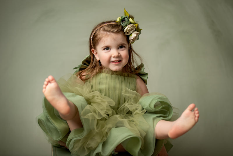 A young girl stands barefoot on a green box wearing a light olive green tulle dress with layered ruffles and oversized bow-tied shoulder straps, posing against a matching green backdrop.

