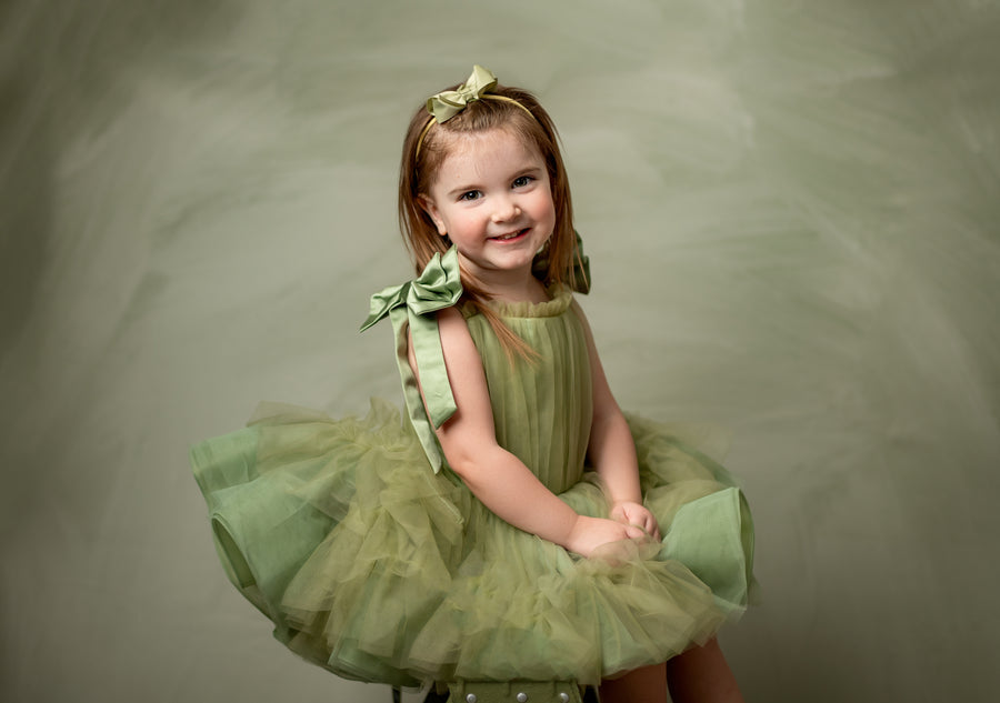 Toddler girl in a whimsical green dress smiles softly while standing on a box, styled with a floral headband and soft pleated tulle skirt.

