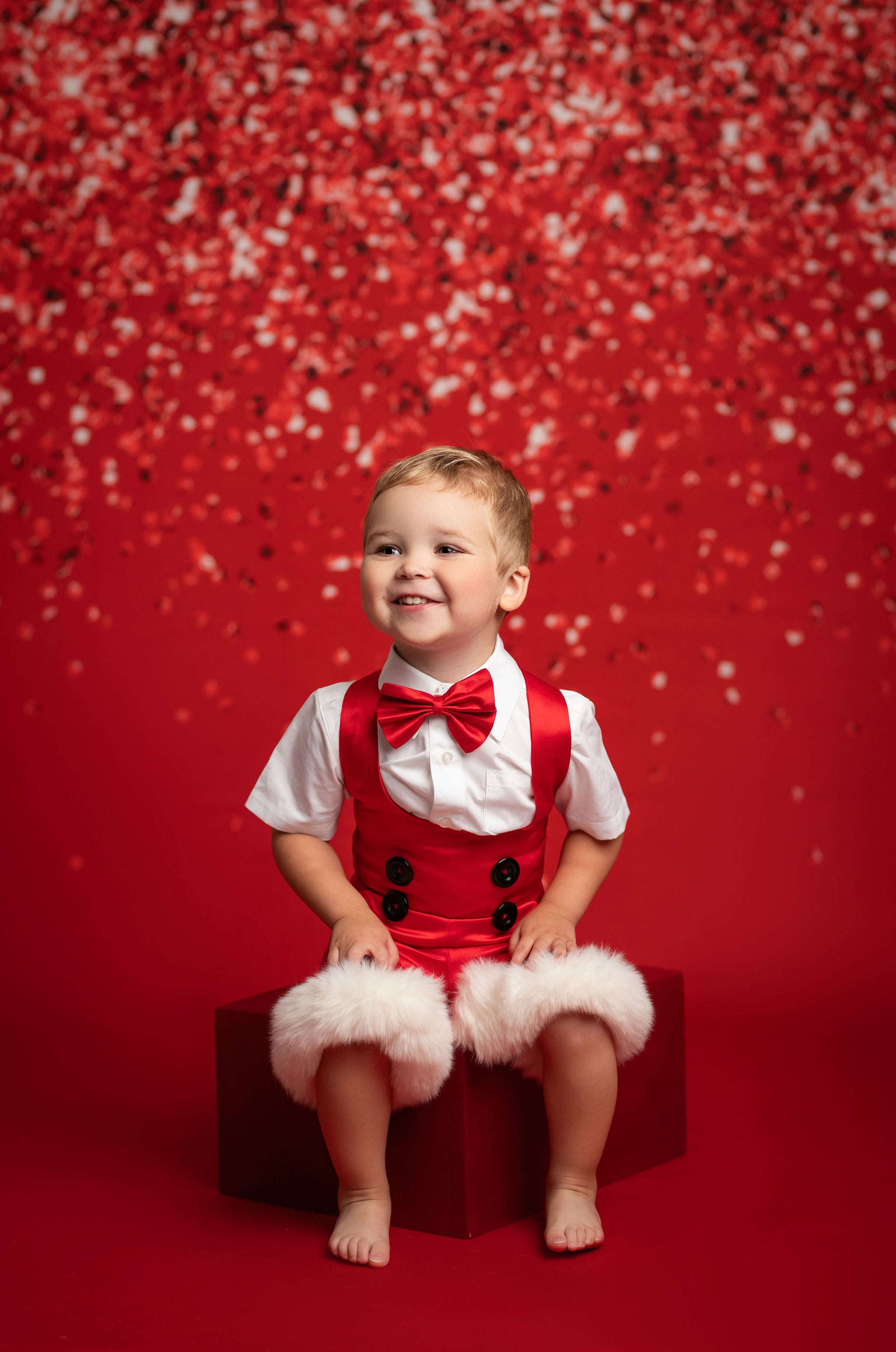 Child in a red outfit with suspenders and a bow tie sitting on a red box against a red background with white polka dots.