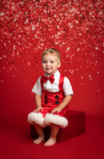 Child in a red outfit with a bow tie sitting on a red box against a red background with confetti.