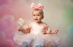 Baby in a pink dress with a large bow and a colorful lollipop against a soft, multicolored background.