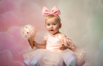 Baby in a pink dress with a large bow holding a decorative ice cream cone against a soft, blurred background.