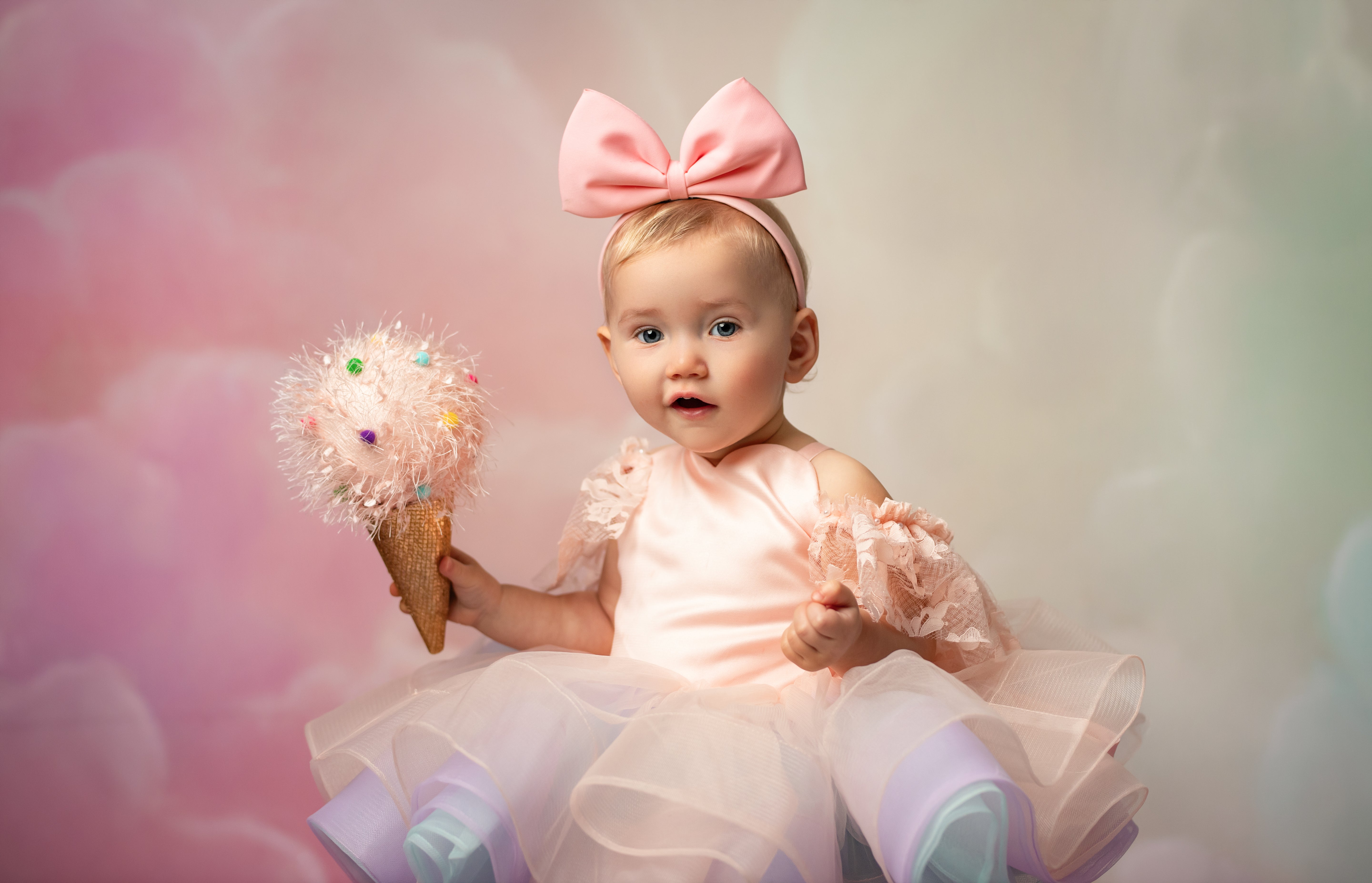 Baby in a pink dress with a large bow holding a decorative ice cream cone against a soft, blurred background.