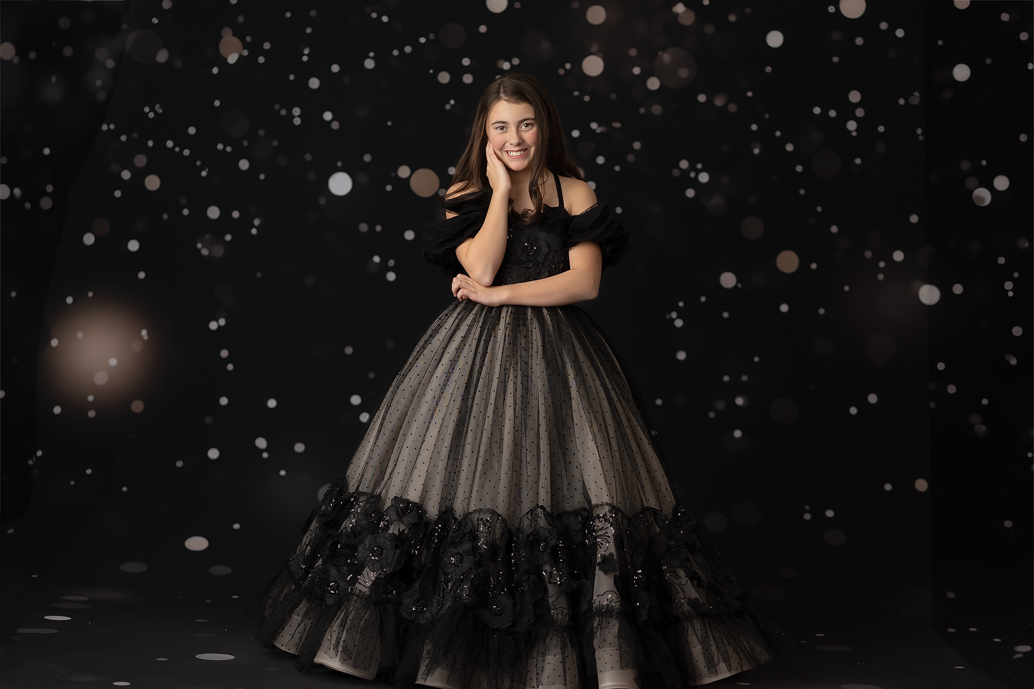 Young woman in a black couture gown with ruffled off-shoulder sleeves, posing against a sparkling bokeh background.