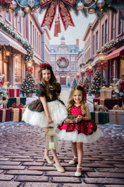 Two young girls in festive Christmas dresses standing on a cobblestone street backdrop decorated with trees, ornaments, and gift boxes. The older girl wears a red and green plaid dress with a white tulle skirt and holds a lantern, while the younger girl wears a red and black polka dot dress with a white tulle skirt and holds a small basket.
