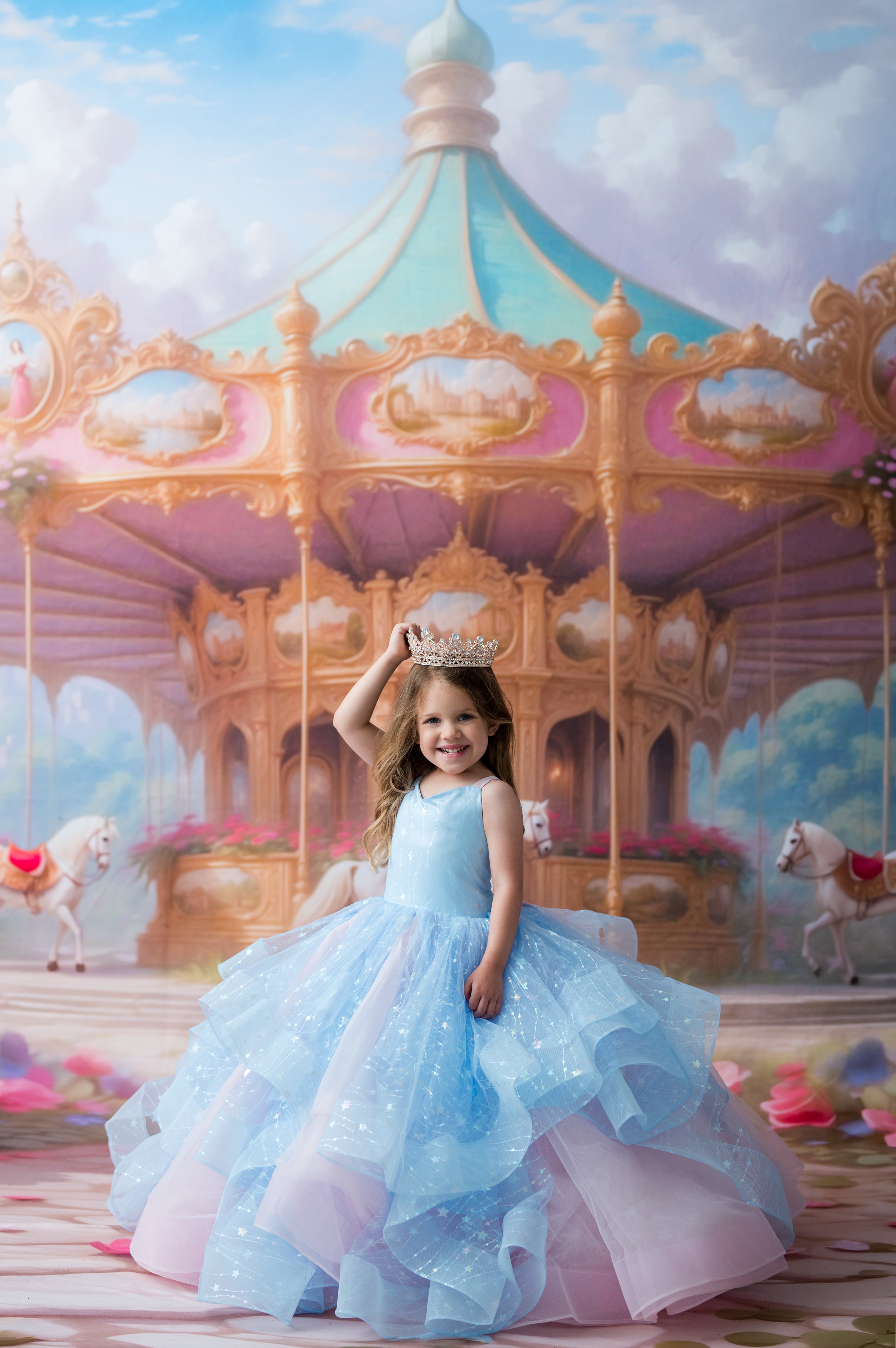 A young girl in a sparkling light blue and lavender ball gown with a gold tiara stands in front of a painted carousel backdrop, posing with one hand raised to her tiara.