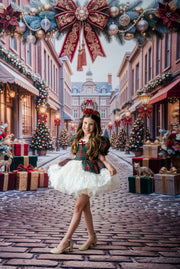 A young girl stands in a festive Christmas town street wearing a plaid and white tulle holiday dress with a matching headpiece. The background features decorated storefronts, Christmas trees, and a large red bow hanging overhead.