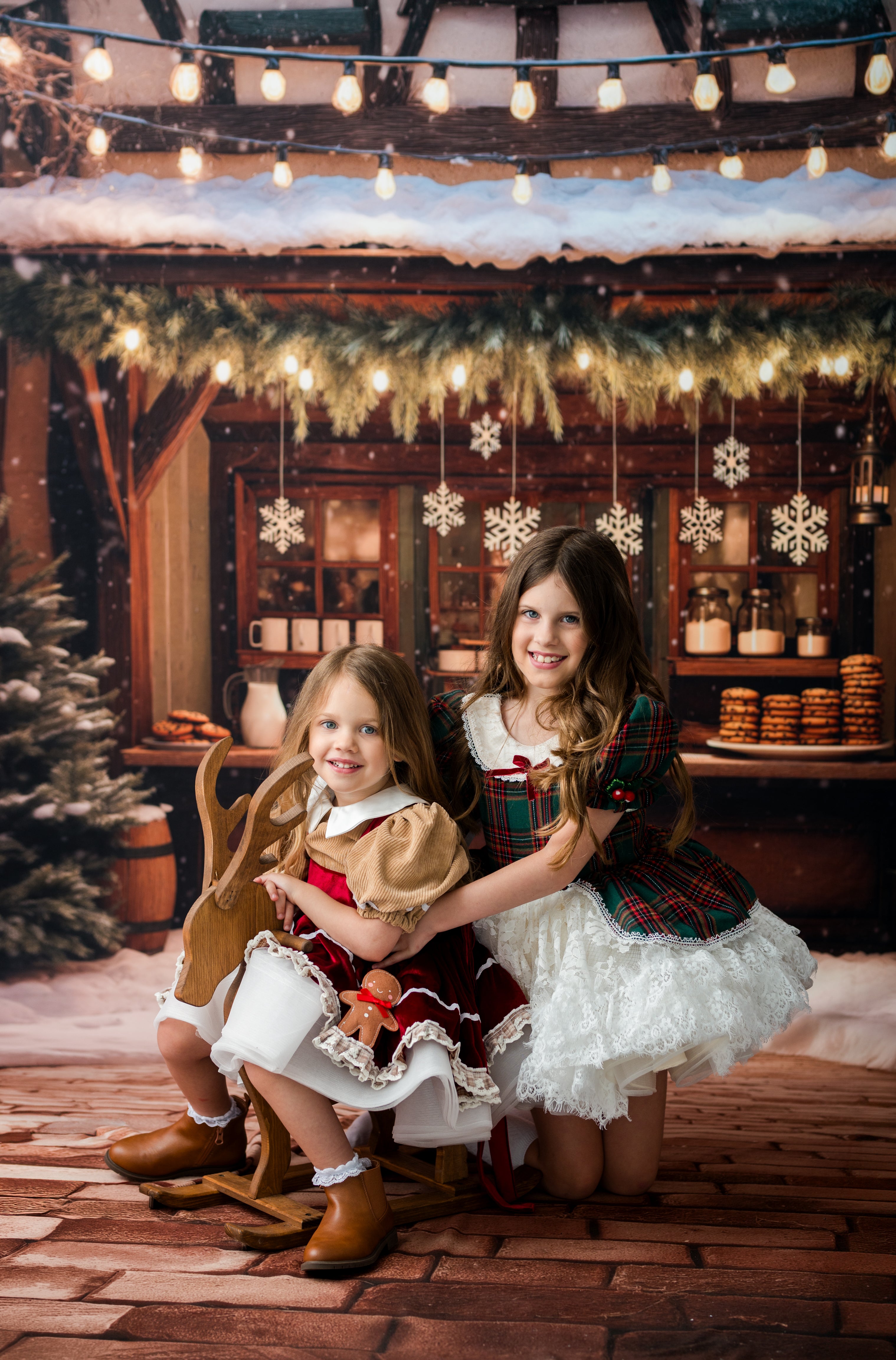 Two young girls in festive Christmas dresses posing with a wooden sleigh on a rustic holiday set with warm lights, snowflakes, and gingerbread decorations.