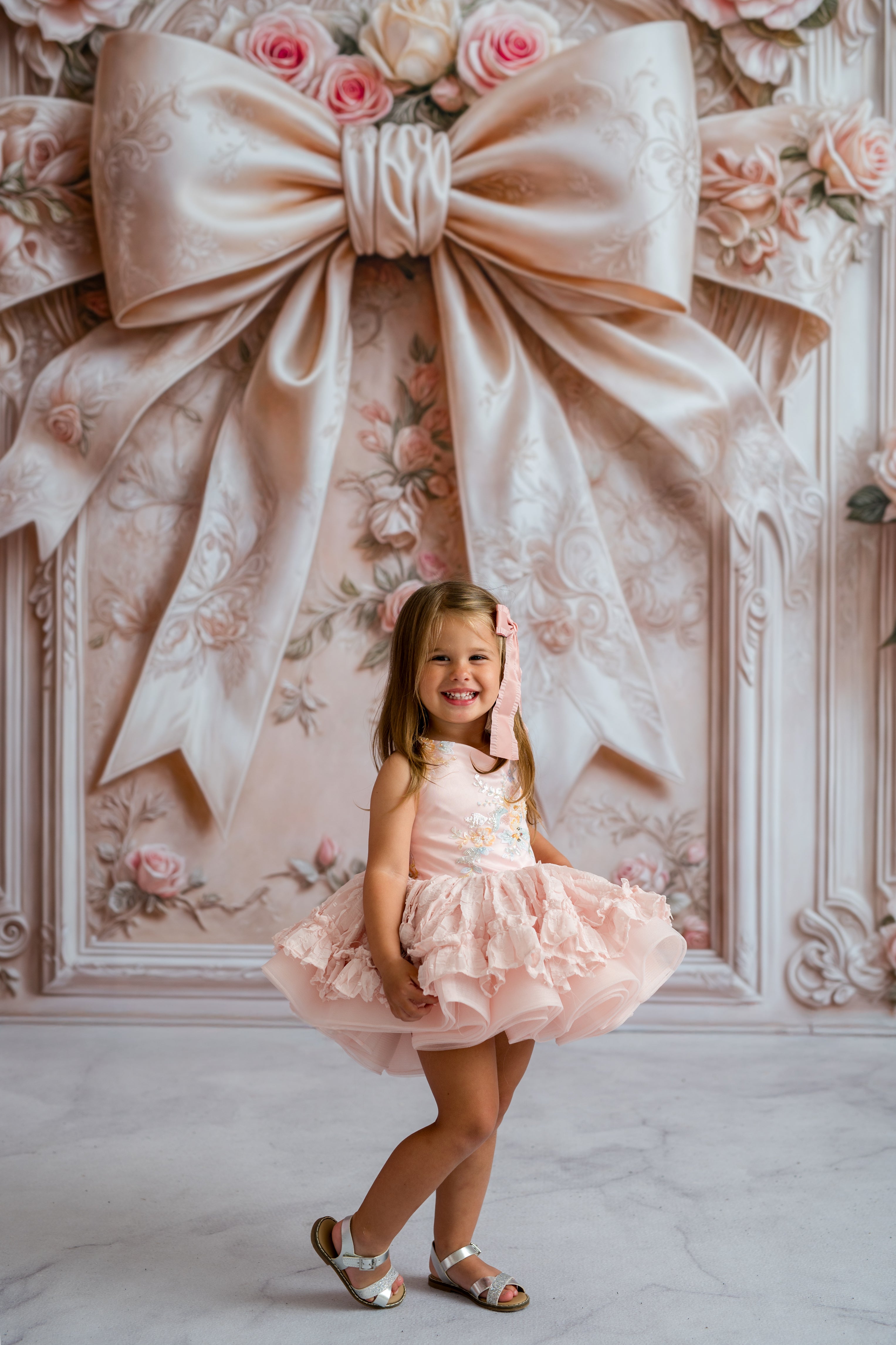Young girl in a pink dress standing in front of a decorative wall with a large bow and floral patterns.