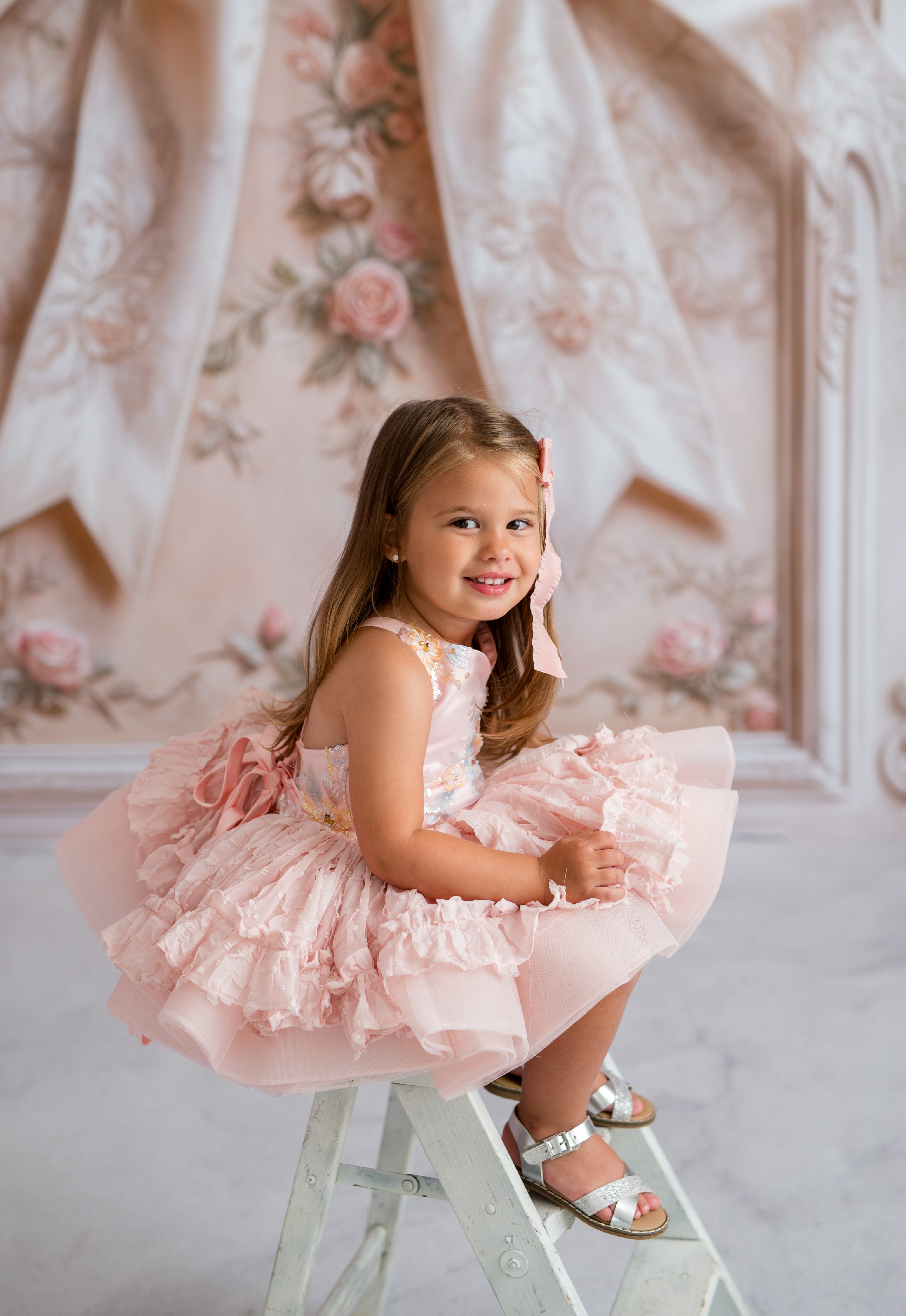 Young girl in a pink dress sitting on a ladder with floral backdrop