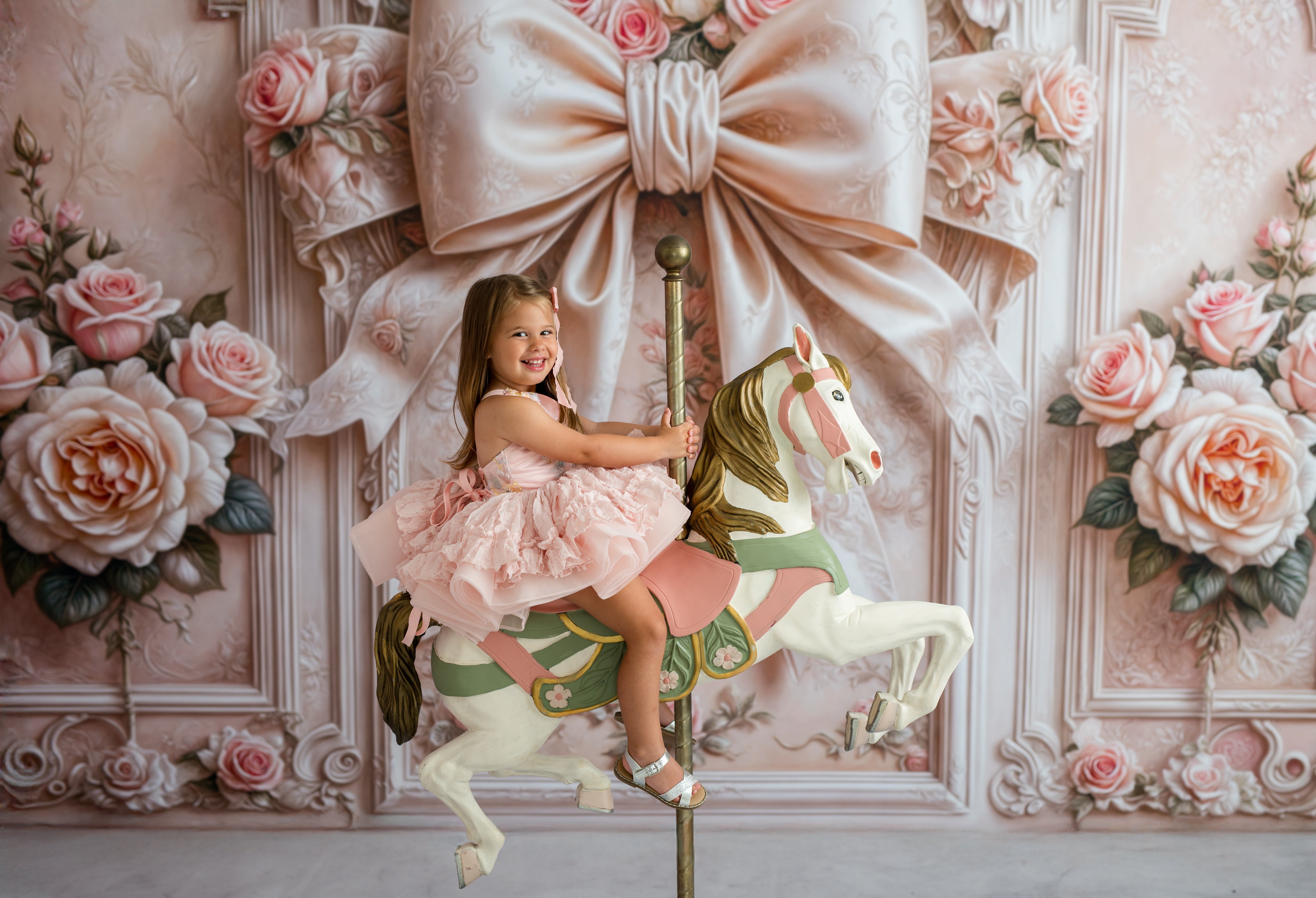 Young girl in a pink dress sitting on a carousel horse against a decorative wall with flowers and a large bow.