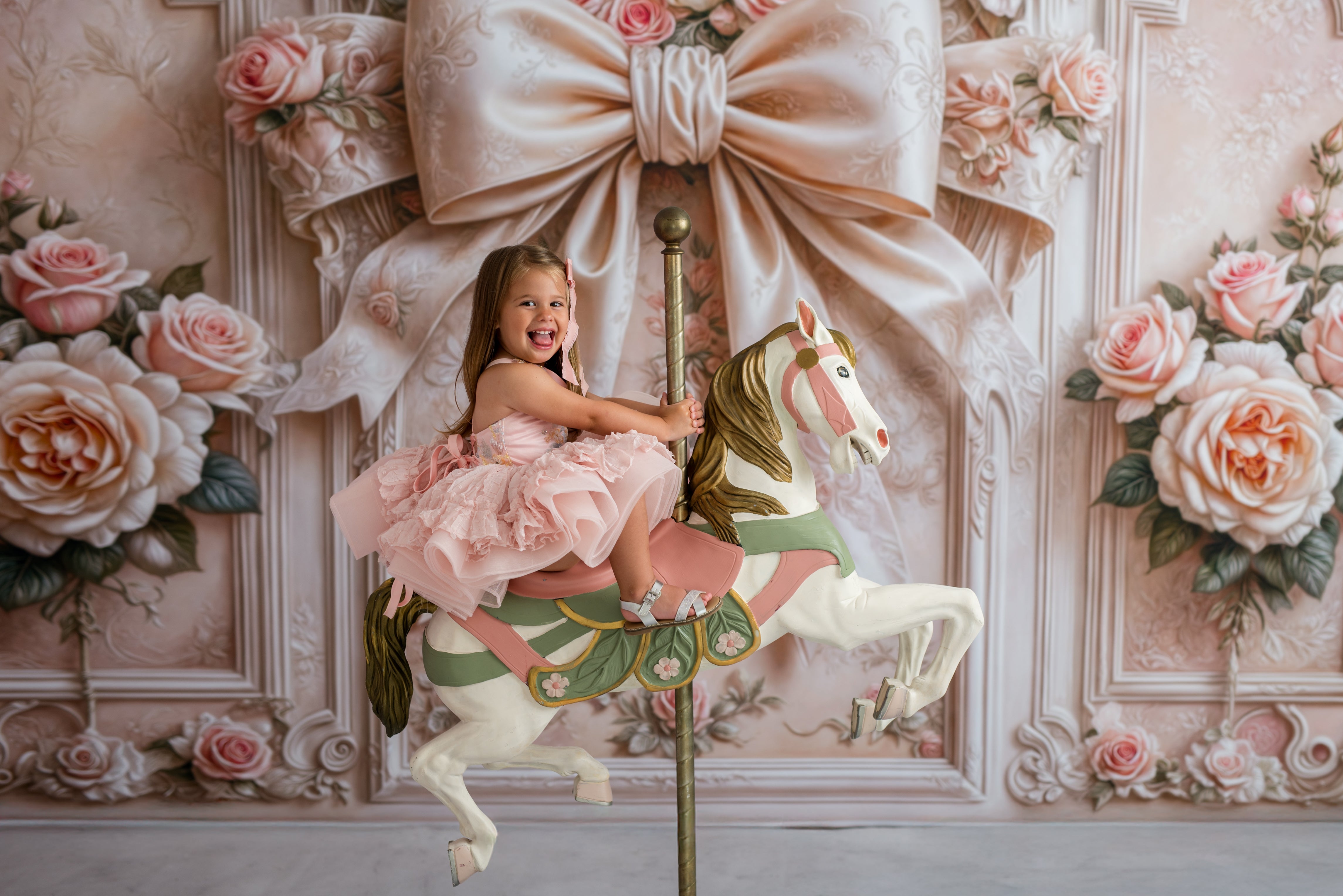 Young girl in a pink dress riding a carousel horse against a decorative floral and bow backdrop.