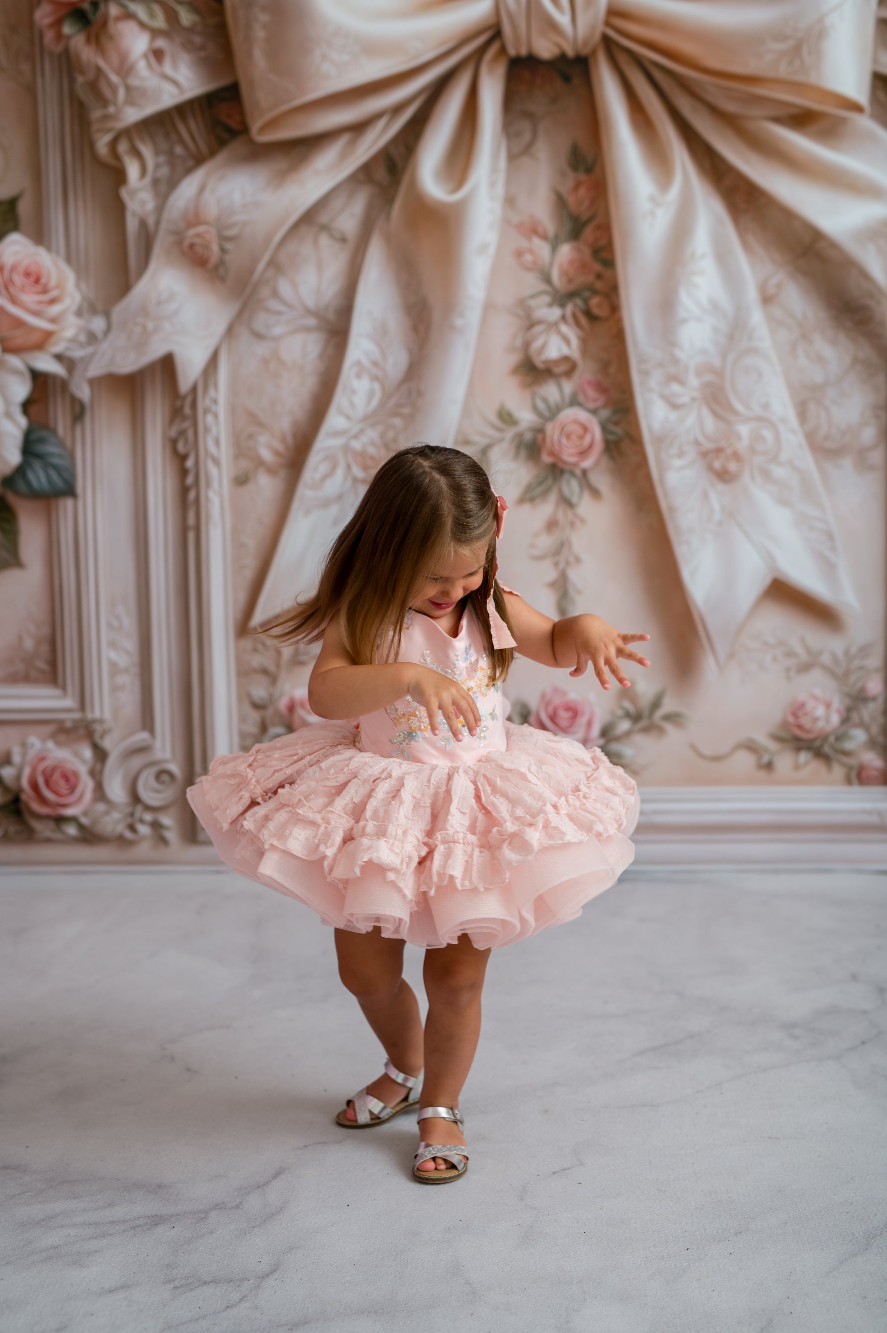 Young girl in a pink dress standing in a room with decorative wallpaper and floral decorations.