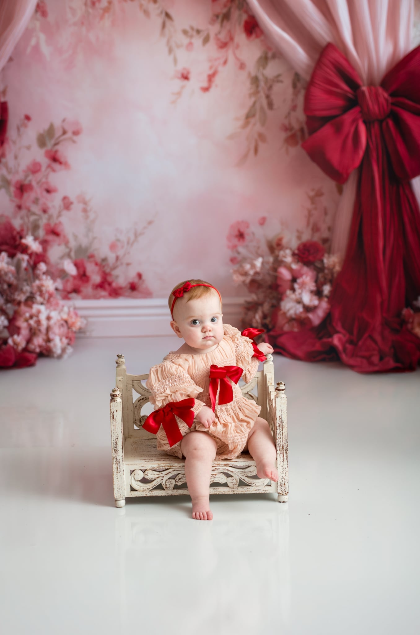 Baby in a pink outfit with red bows sitting on a small bed against a floral backdrop.