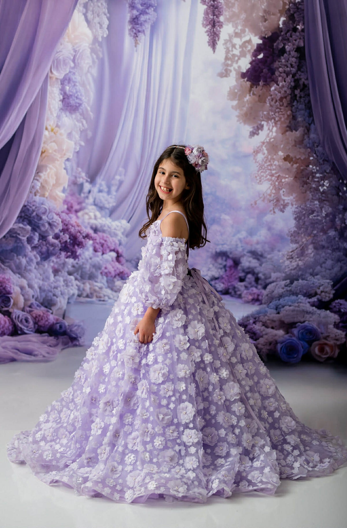 Young girl in a floral dress standing in front of a decorative floral and curtain backdrop.
