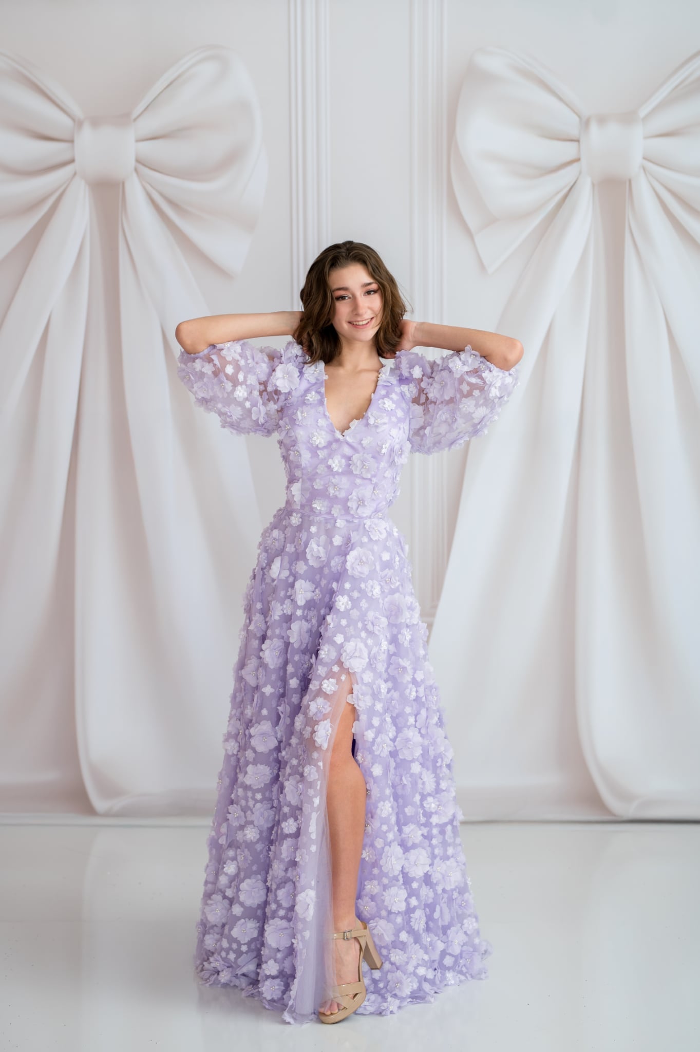Woman in a purple floral dress posing against a white curtain with large bows.