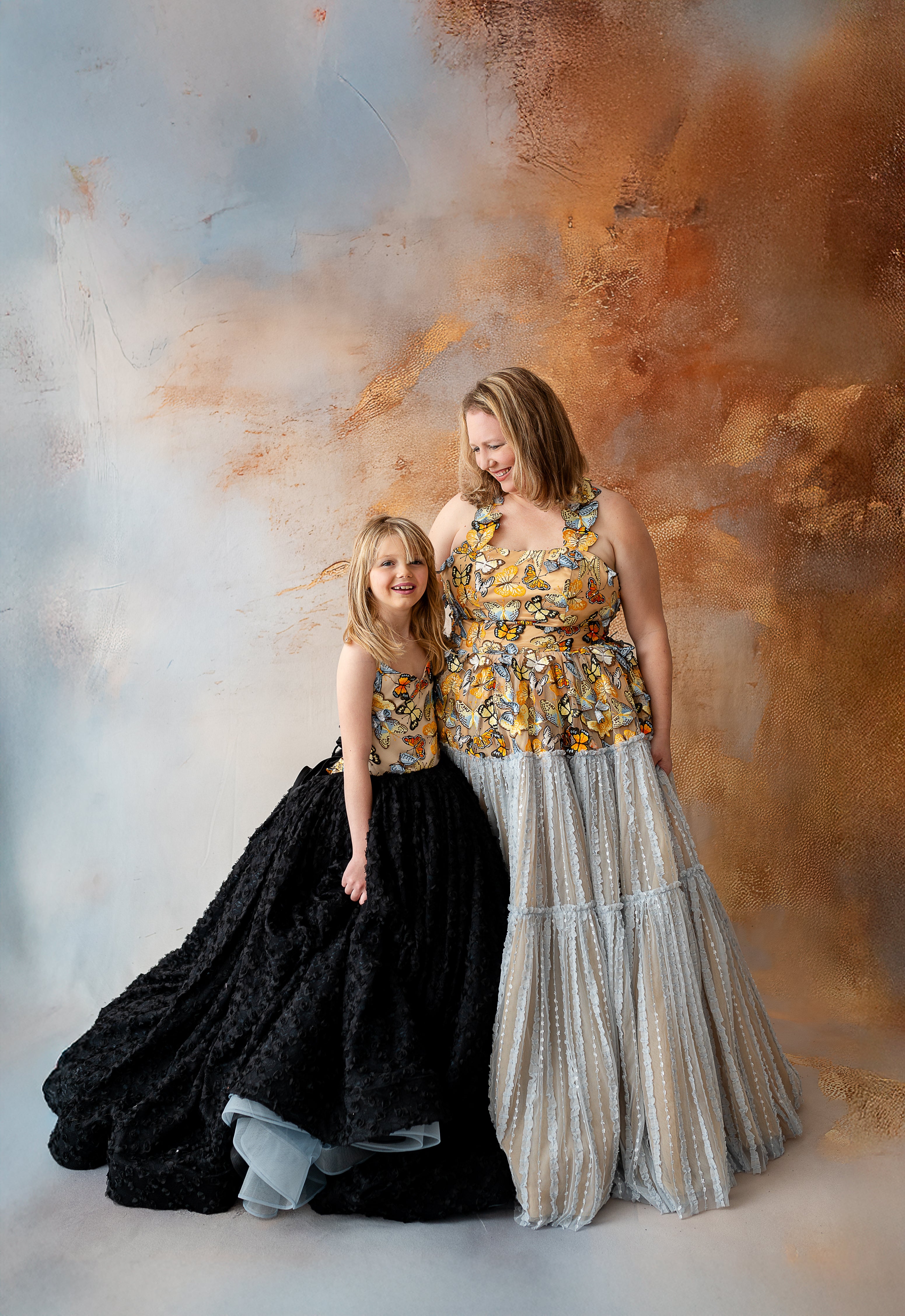 Mom and daughter share a tender moment, dressed in coordinating gowns adorned with delicate butterfly motifs. The soft, flowing fabric of their dresses creates a harmonious, elegant look as they embrace the beauty of the moment together.