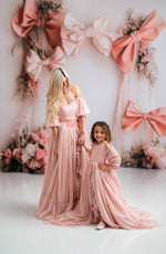 Two women in matching pink dresses standing in front of a decorative backdrop with large pink bows and flowers.