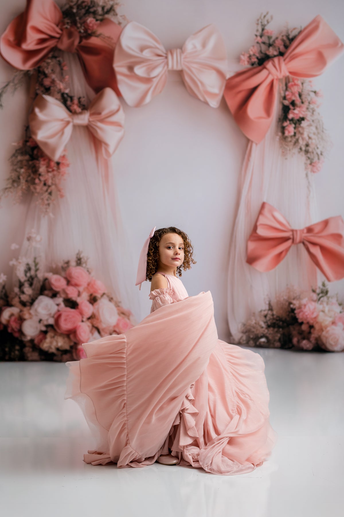 Young girl in a pink dress sitting in front of decorative bows and flowers on a white wall.