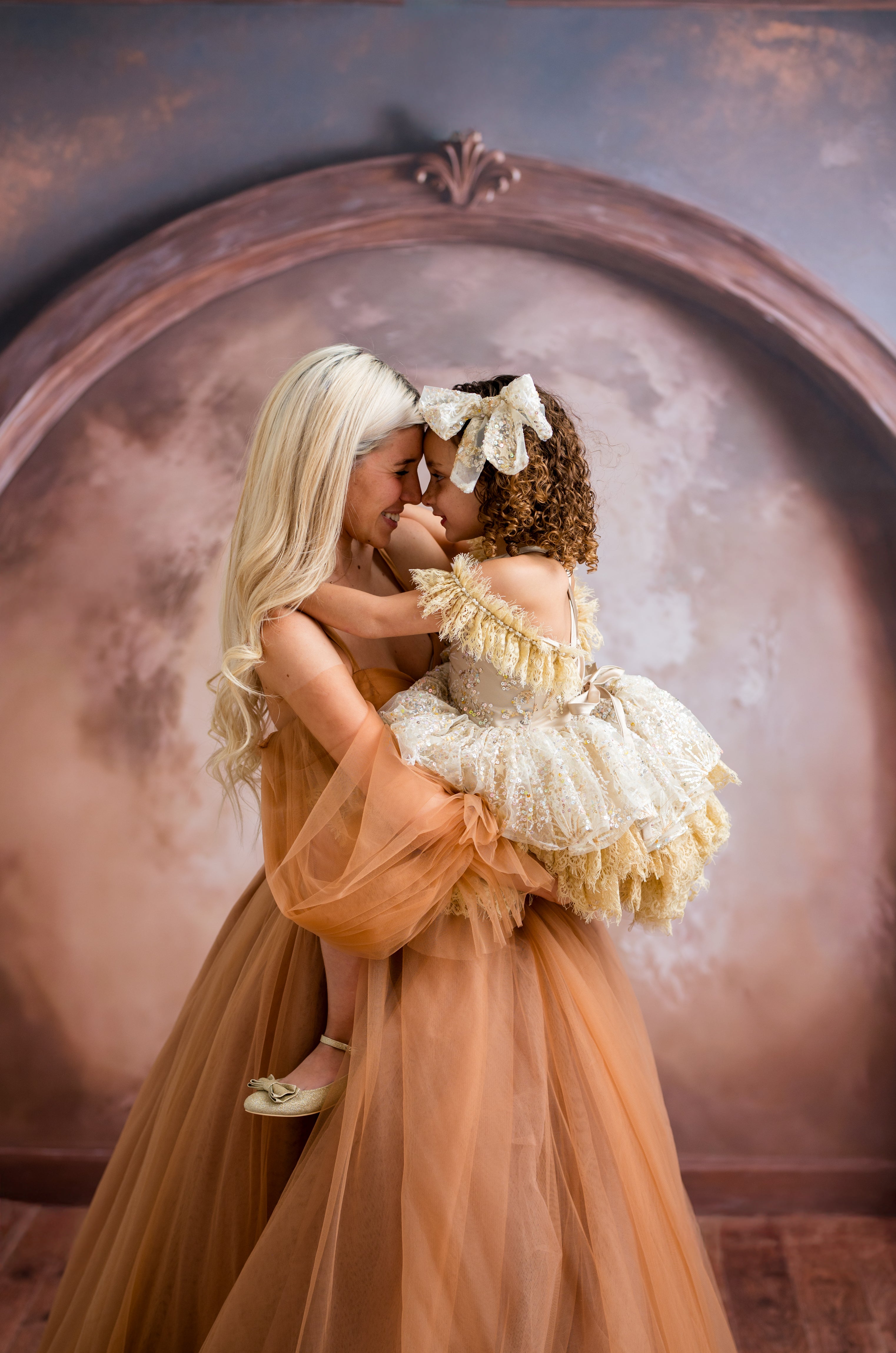 Two women in elegant dresses embracing in front of a decorative mirror.