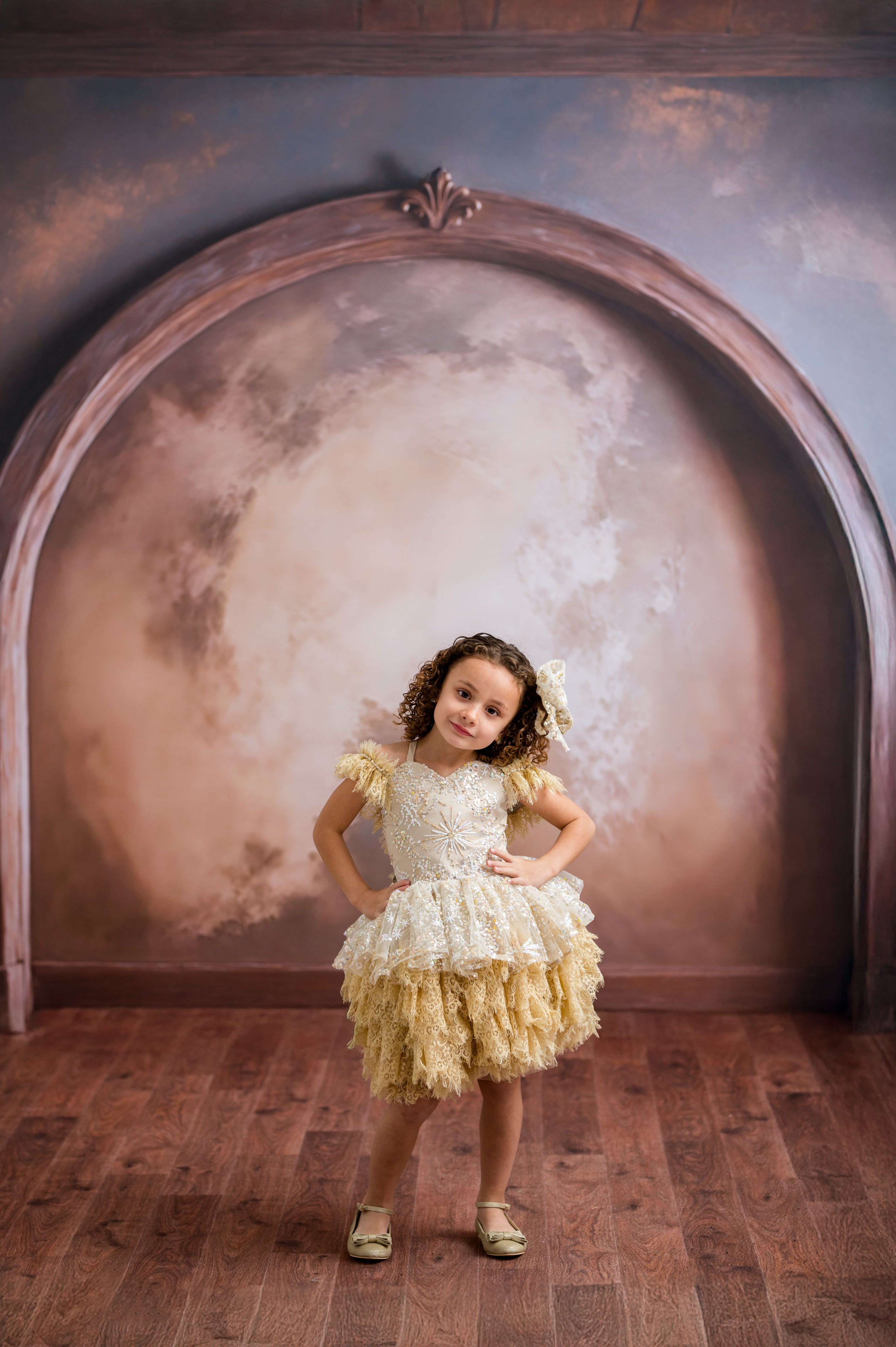 Young girl in a yellow dress standing in front of an ornate mirror with a textured wall background.