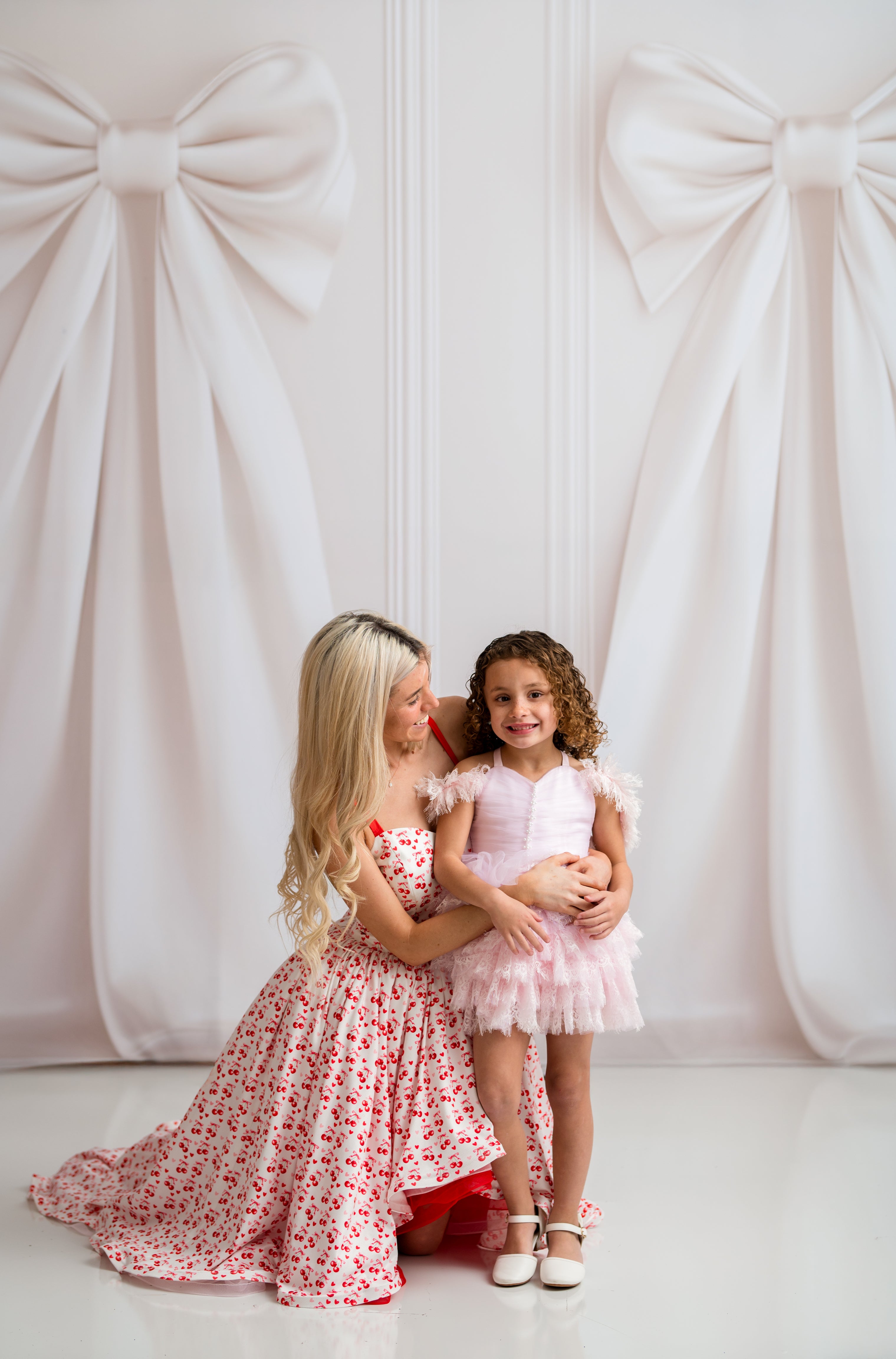 Two women in floral dresses standing in front of a white wall with large decorative bows.