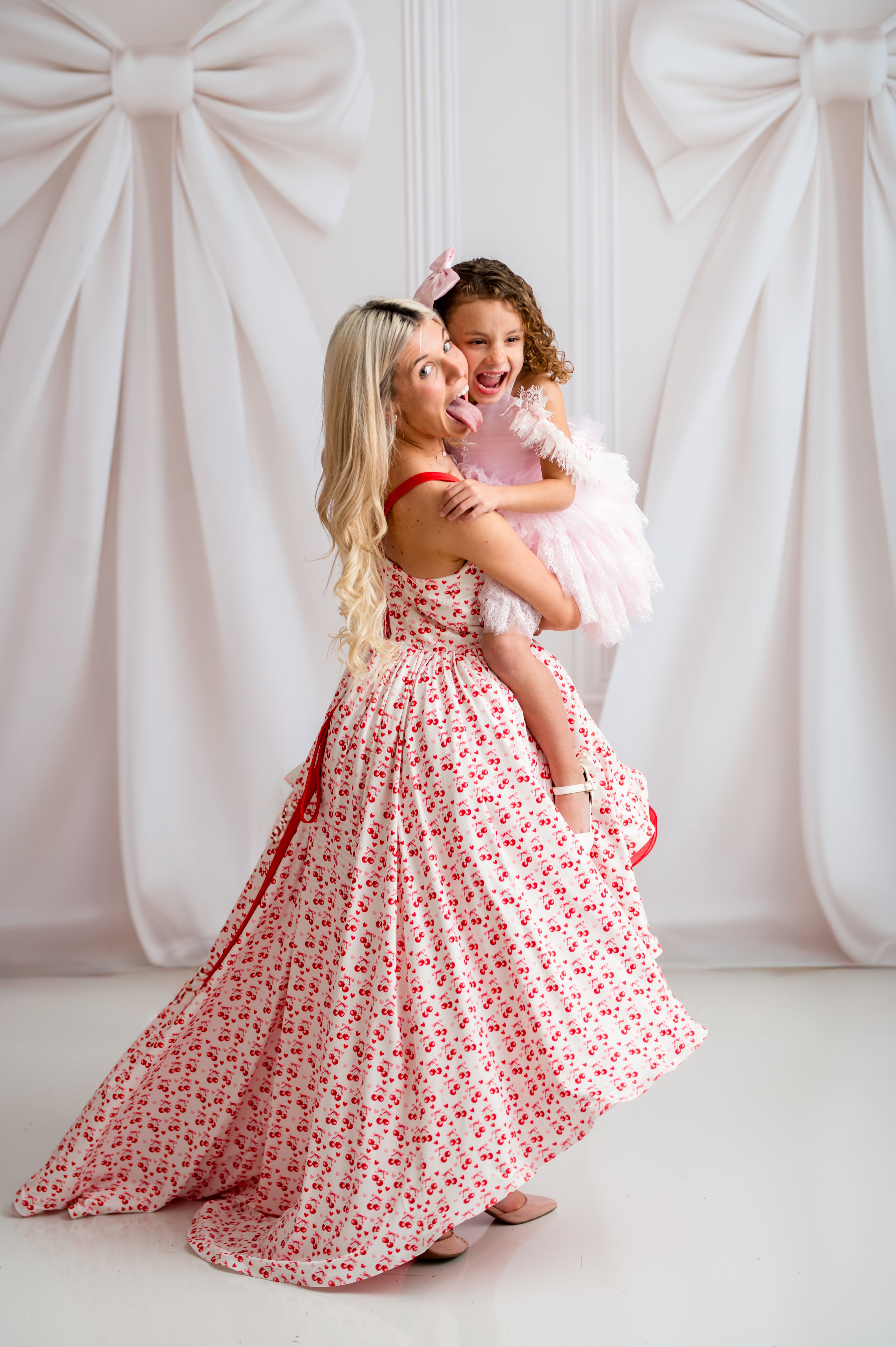 Woman in a floral dress holding a child against a white backdrop with large decorative bows.