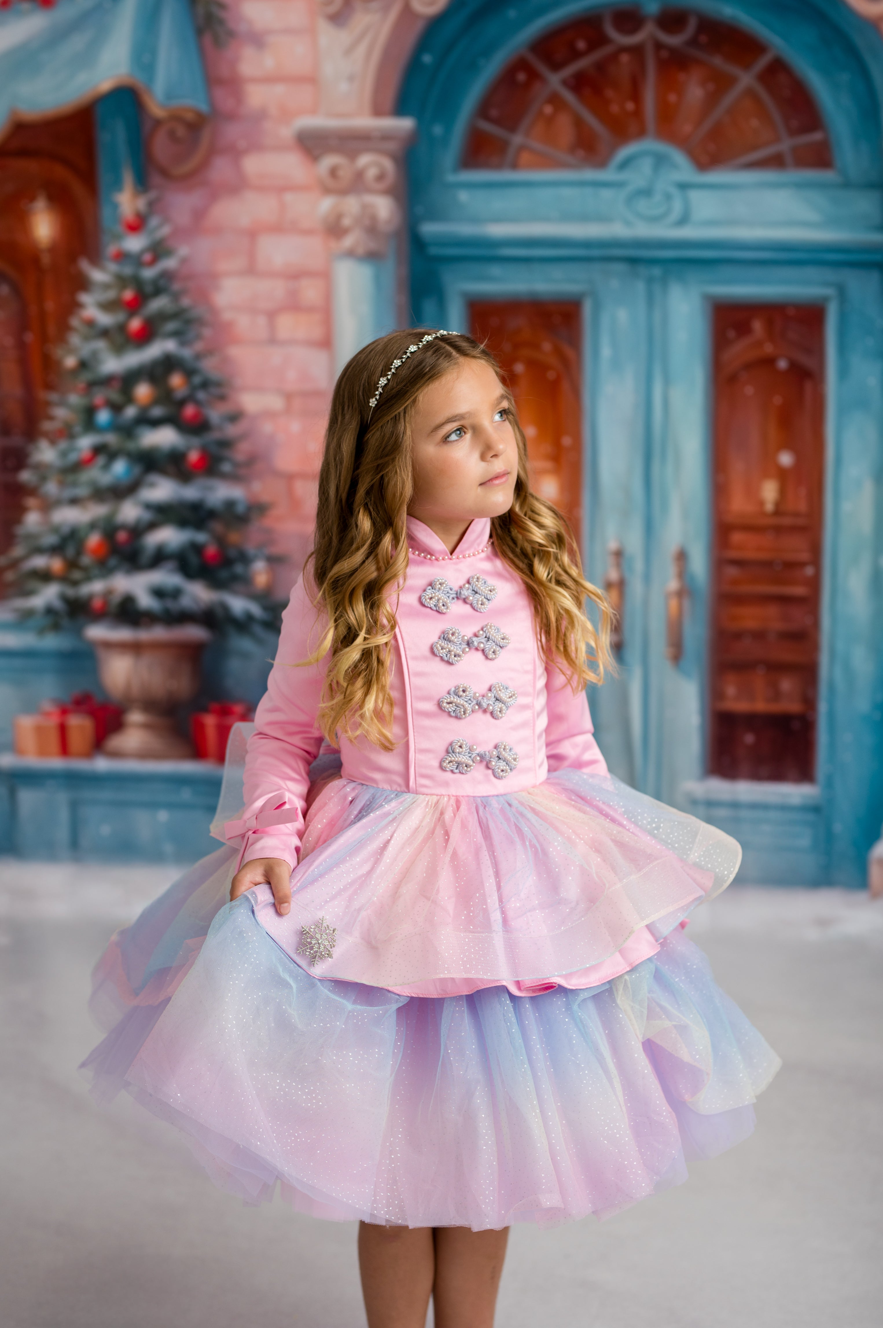 Young girl in a pink dress standing in front of a festive background with a Christmas tree and blue door.