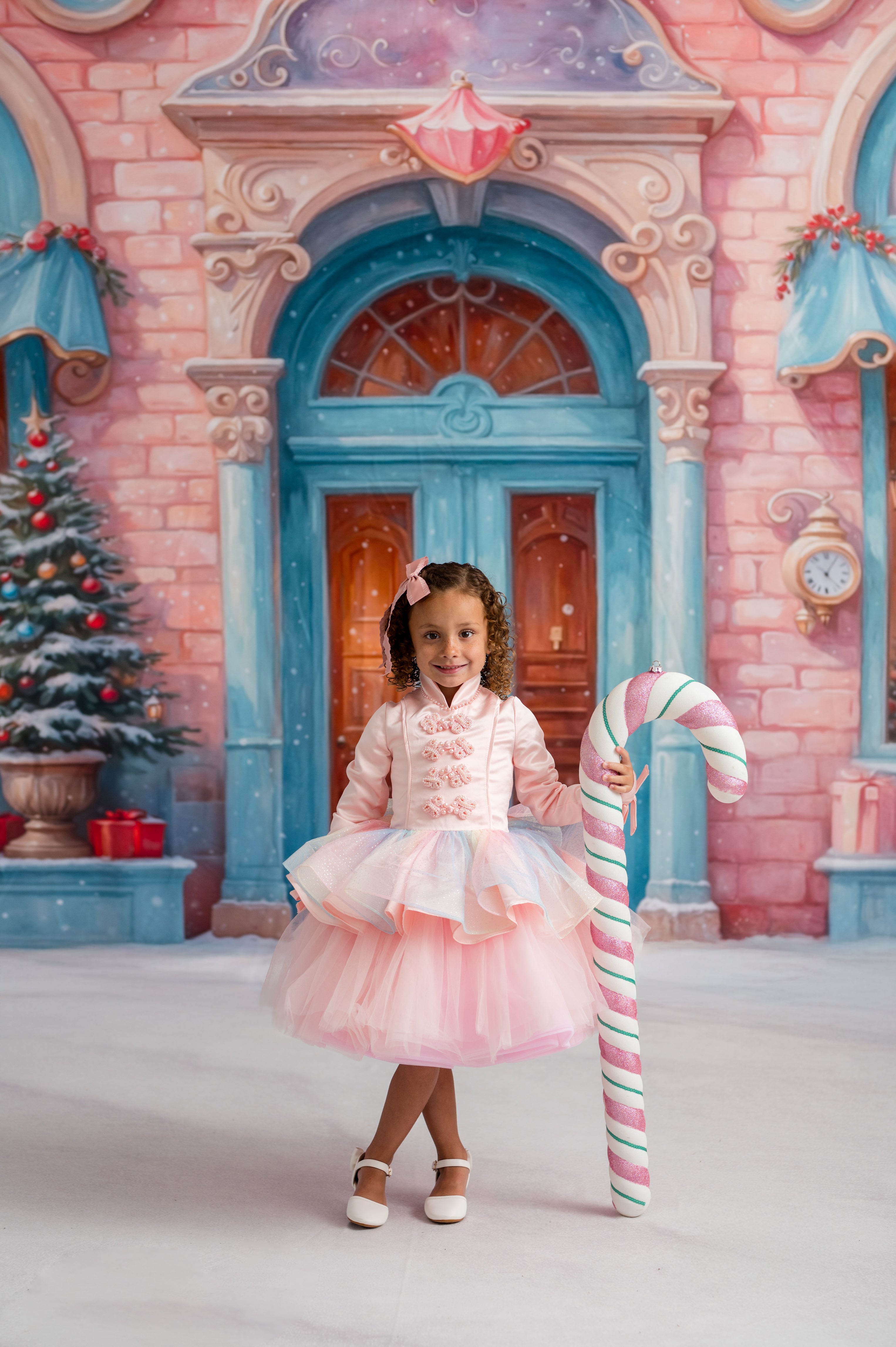Young girl in a pink dress holding a candy cane in front of a festive background with a Christmas tree and candy.