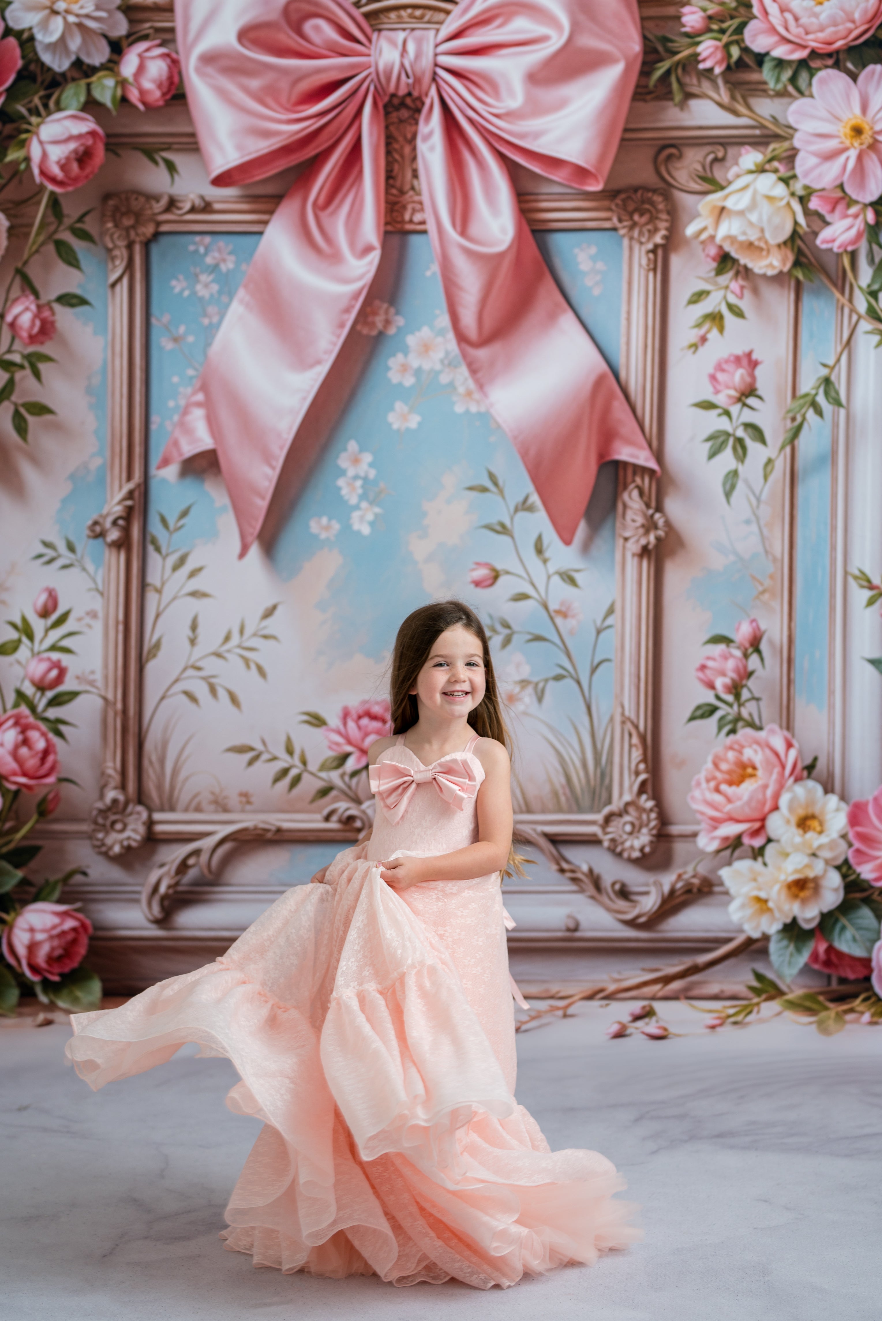 Young girl in a pink dress standing in front of a decorative wall with floral arrangements and a large pink bow.