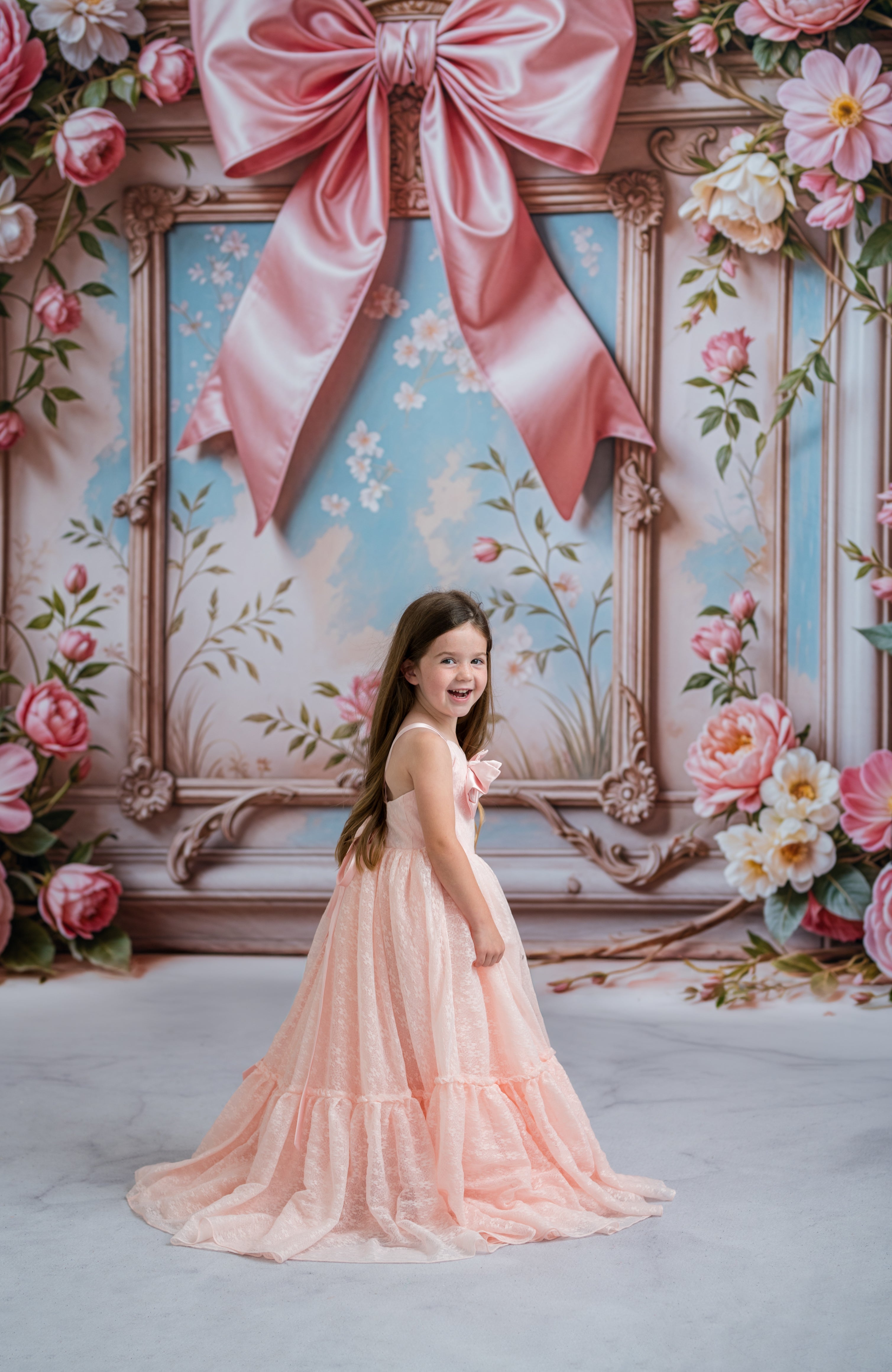 Young girl in a pink dress standing in front of a decorative backdrop with flowers and a large pink bow.