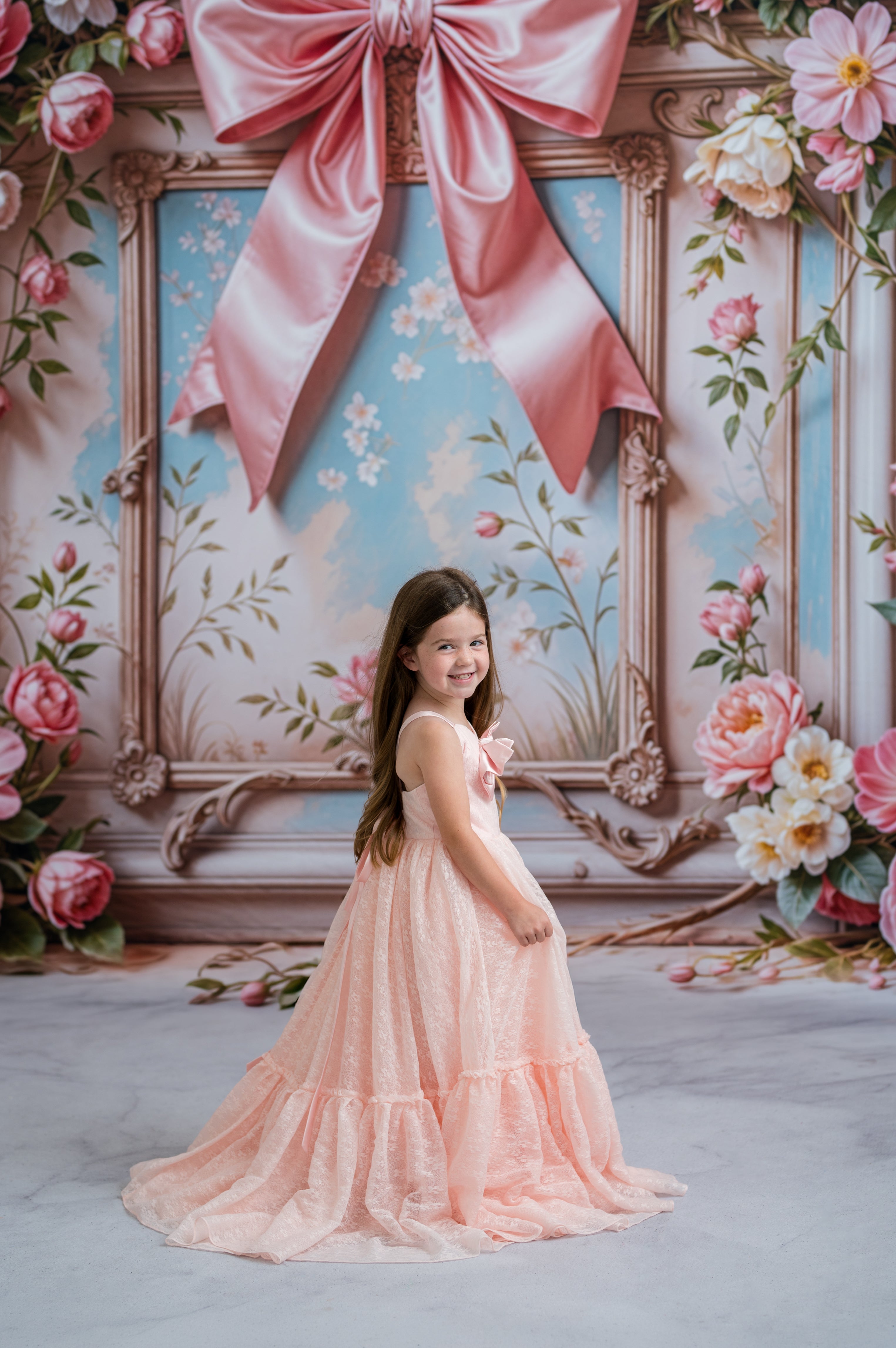 Young girl in a pink dress standing in front of a decorative backdrop with flowers and a large pink bow.