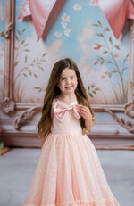 Young girl in a pink dress with a large bow standing in front of a decorative wall.
