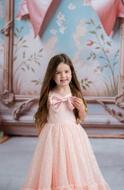 Young girl in a pink dress with a large bow standing in front of a decorative wall.