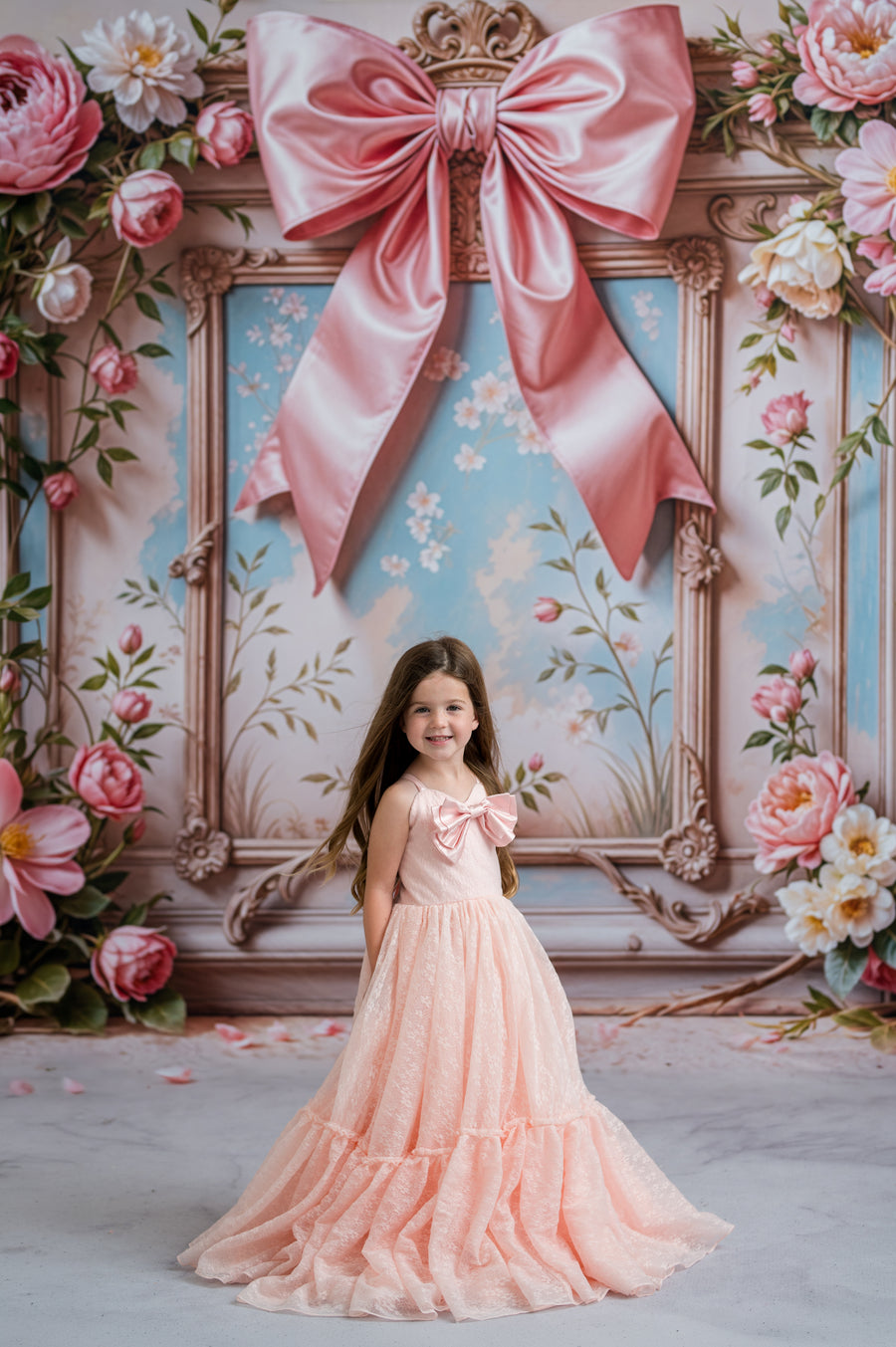 Young girl in a pink dress standing in front of a decorative floral backdrop with a large pink bow.