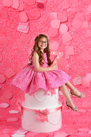 A little girl with glasses and long brown hair celebrates her birthday in a pink sequined dress, posing excitedly beside a large white and pink cake with bows and pearls.

