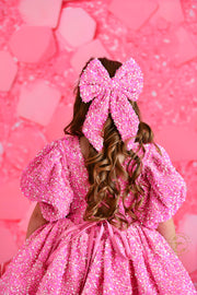 A happy seven-year-old girl stands in a dazzling pink dress, grinning next to a beautifully decorated cake with a glowing "7" on top, against a festive pink background.

