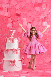 A beaming child in a glittery pink dress twirls with excitement in front of a pink confetti wall, standing next to an elegant, tiered cake adorned with pink bows and a light-up "7" sign.


