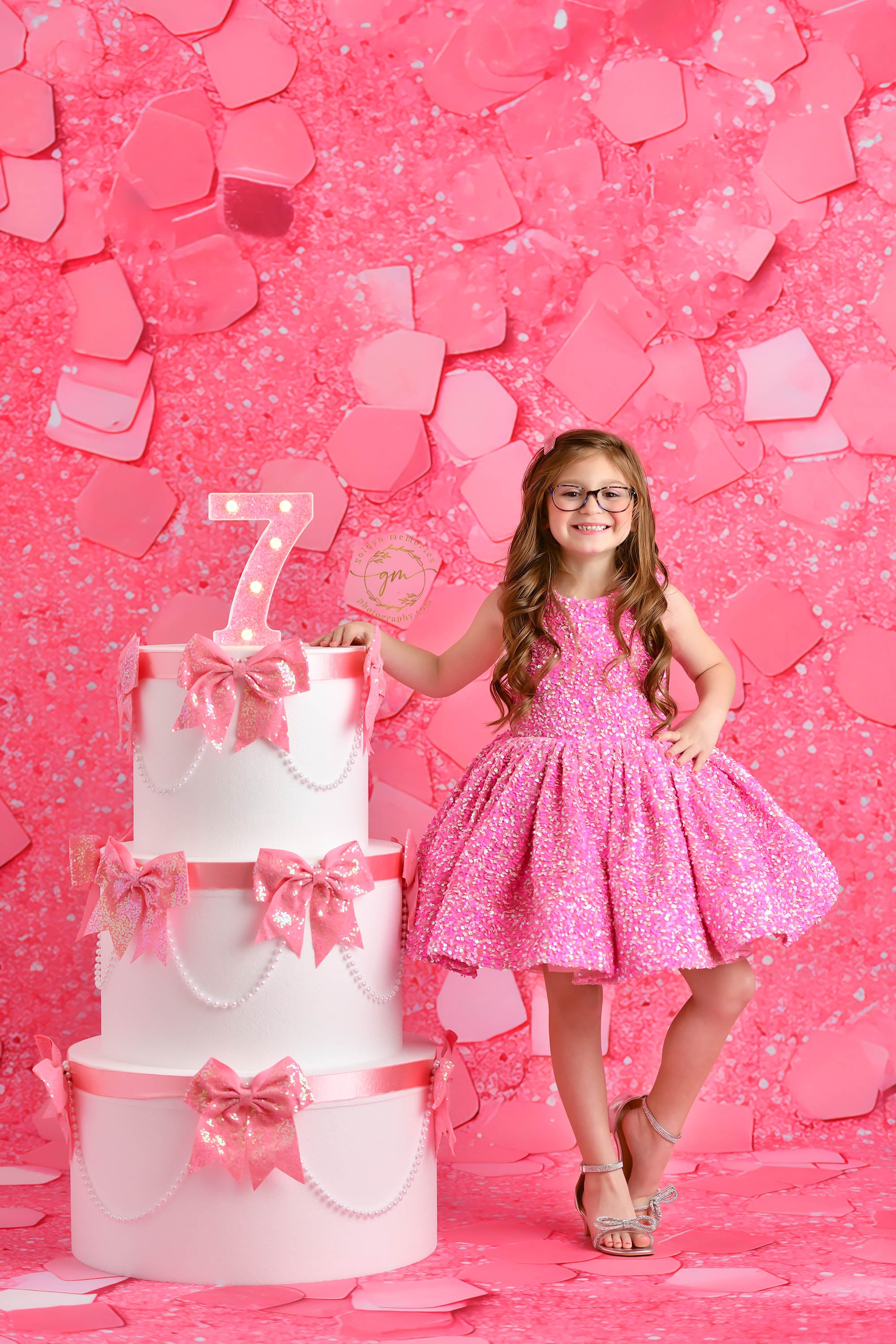 A young birthday girl dressed in a vibrant pink dress with puffed sleeves poses with arms wide open beside a glamorous white cake with pink embellishments, in a pink confetti-filled setting.

