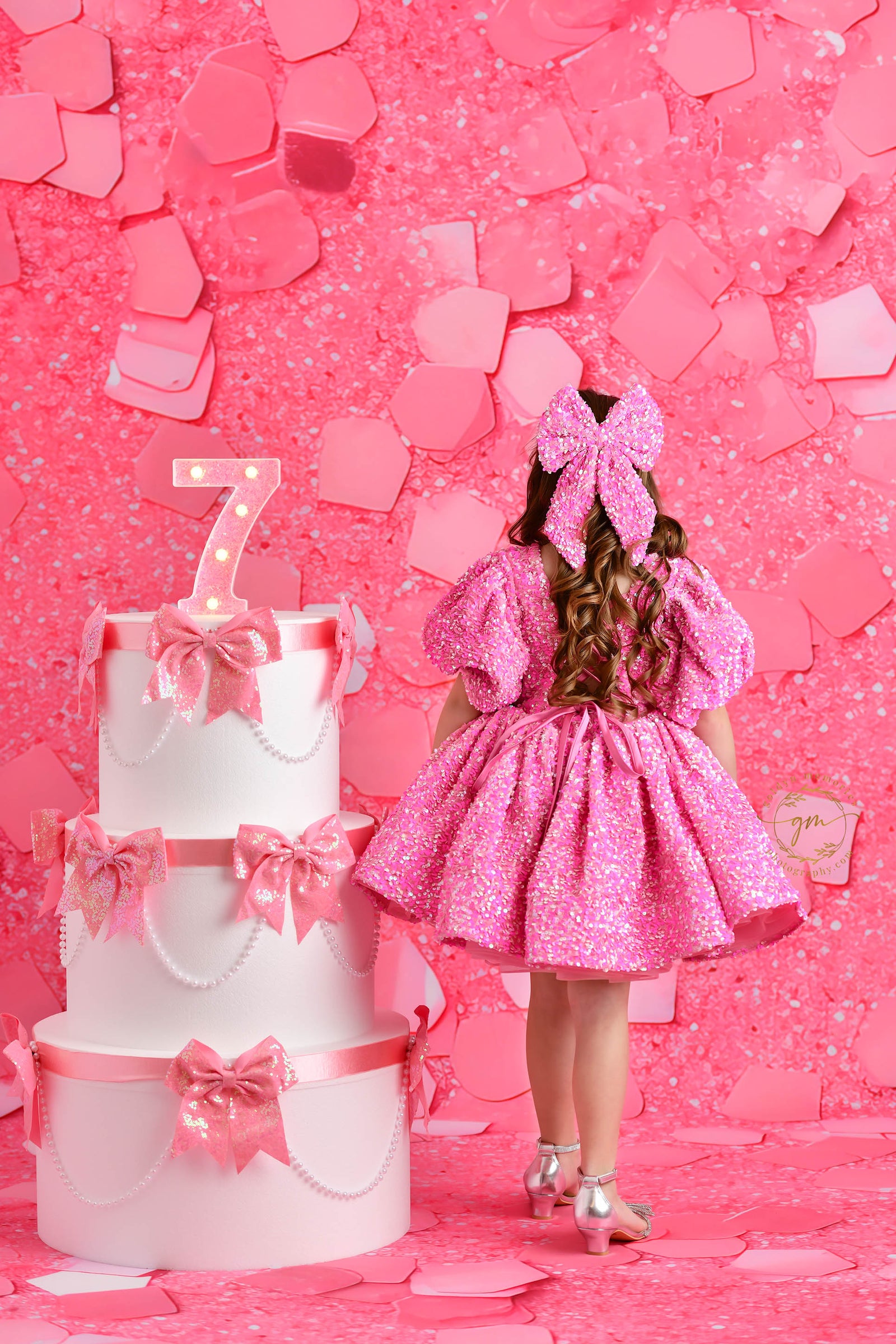 A joyful young girl in a sparkly pink dress with puff sleeves stands with her arms raised in front of a pink confetti-themed backdrop, smiling brightly next to a decorated birthday cake with a glowing number "7" topper.