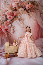 Young girl in a pink floral dress standing next to a doll in a pram, with a floral arch and pink curtains in the background.
