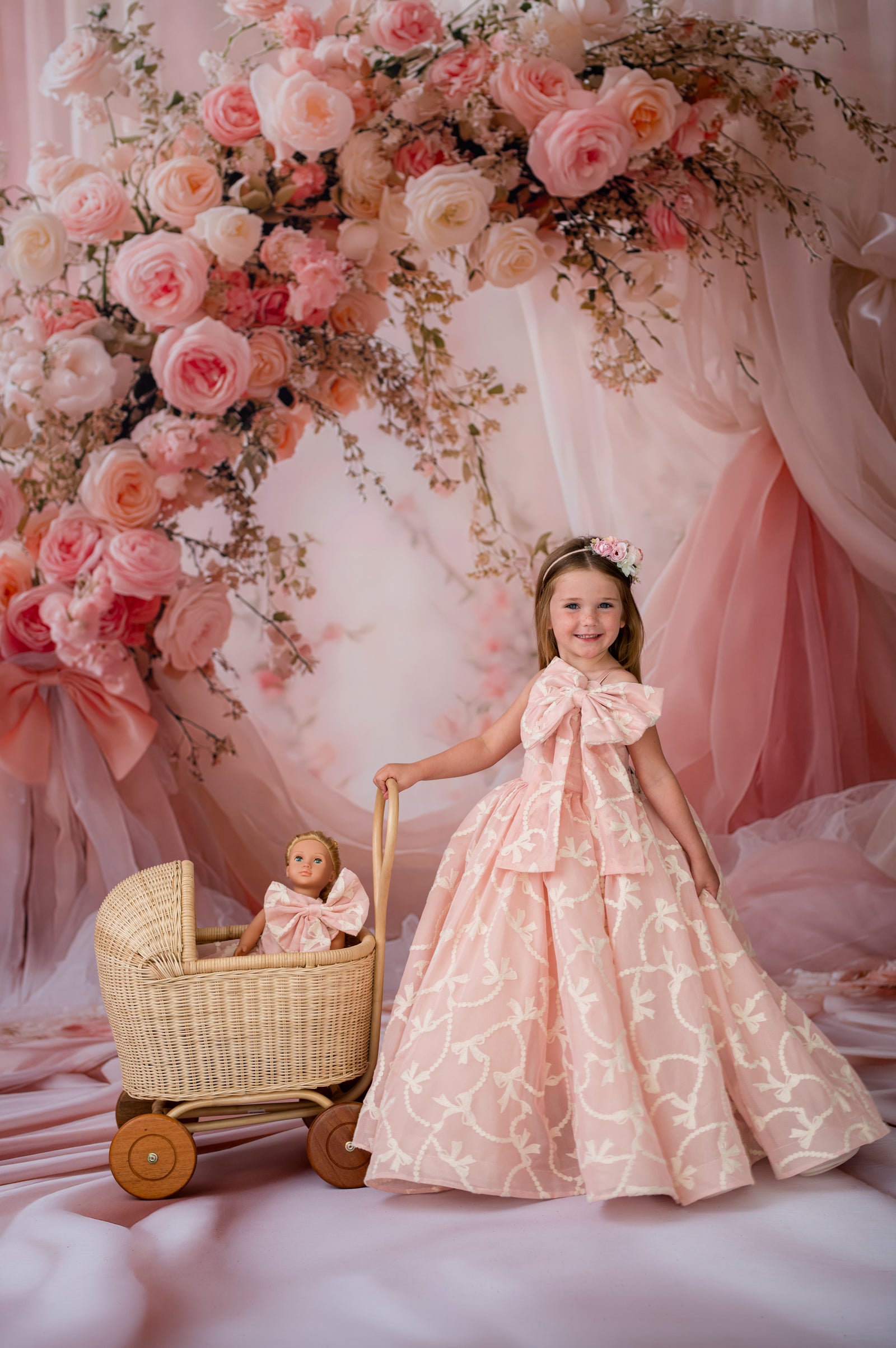 Young girl in a pink floral dress standing next to a doll in a pram, with a floral arch and pink curtains in the background.