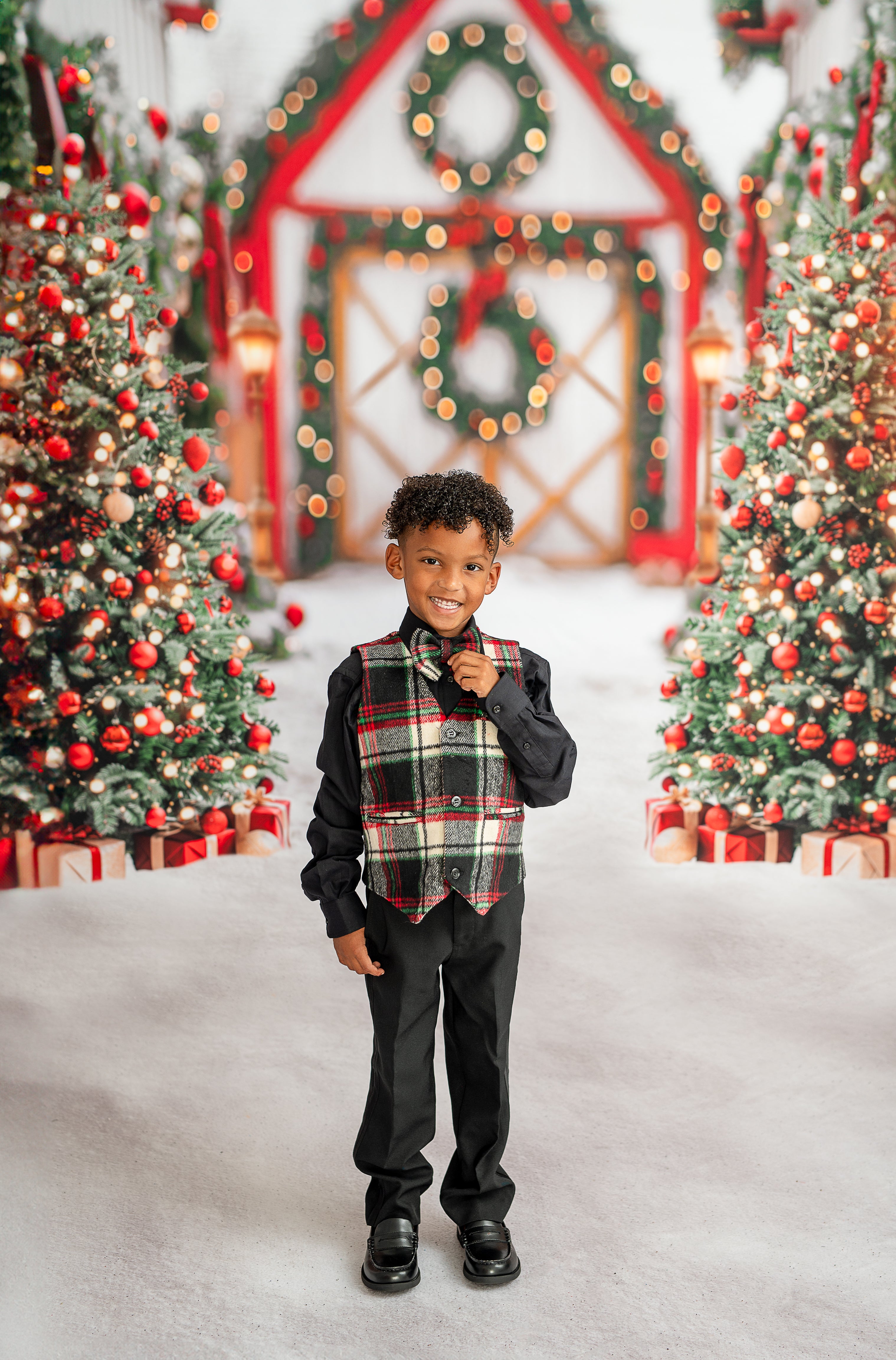 A young boy dressed in a black shirt with a matching plaid vest and shorts holds a green gift adorned with a red bow. The holiday backdrop showcases Christmas trees and a festive doorway with a wreath, creating a joyful seasonal scene.