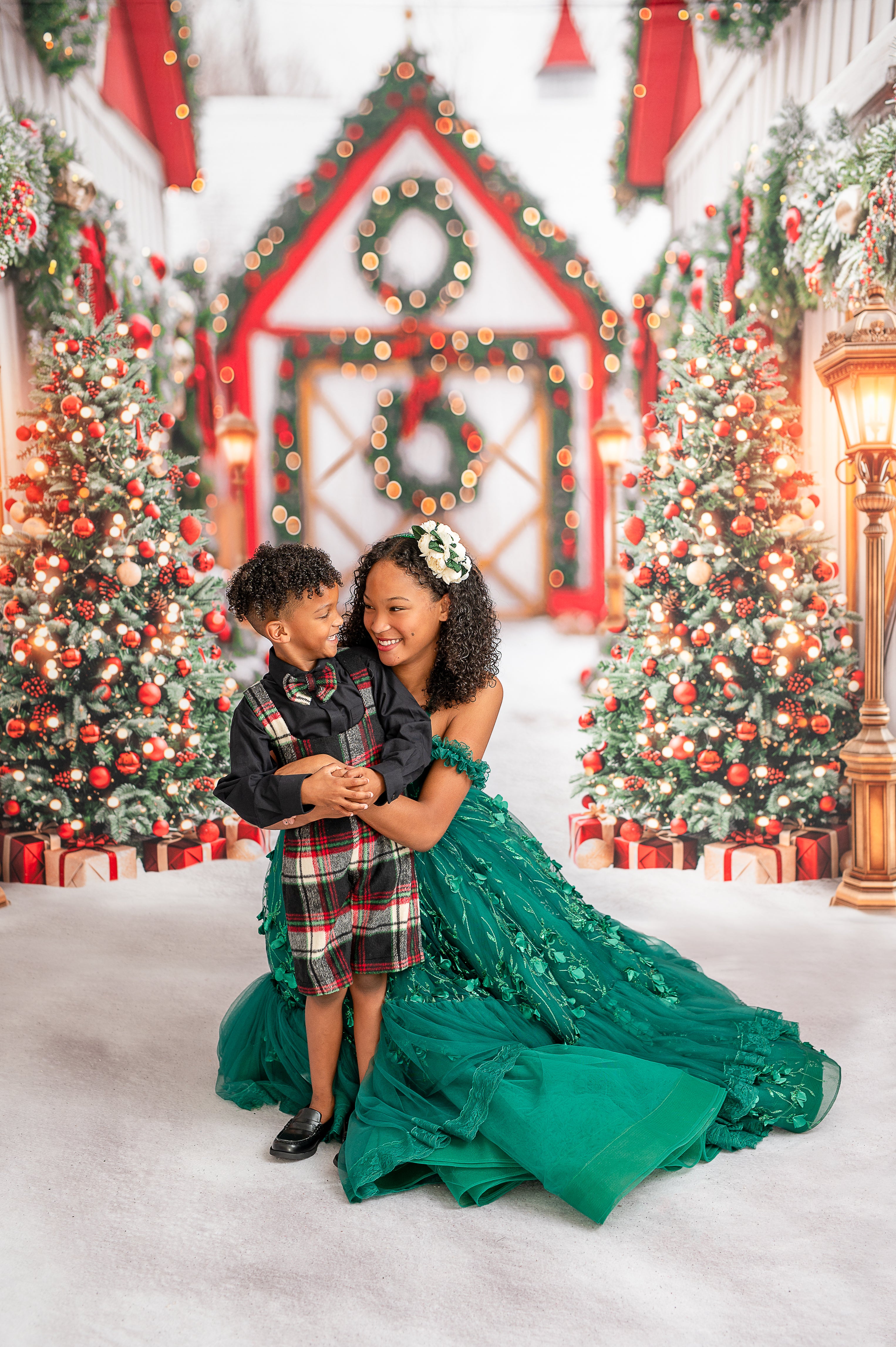 "The image shows a happy boy dressed in a stylish plaid vest and shorts set, complete with a plaid bow tie and black shirt. He holds a wrapped gift, standing in front of an enchanting Christmas setting with decorated trees and a glowing wreath."

