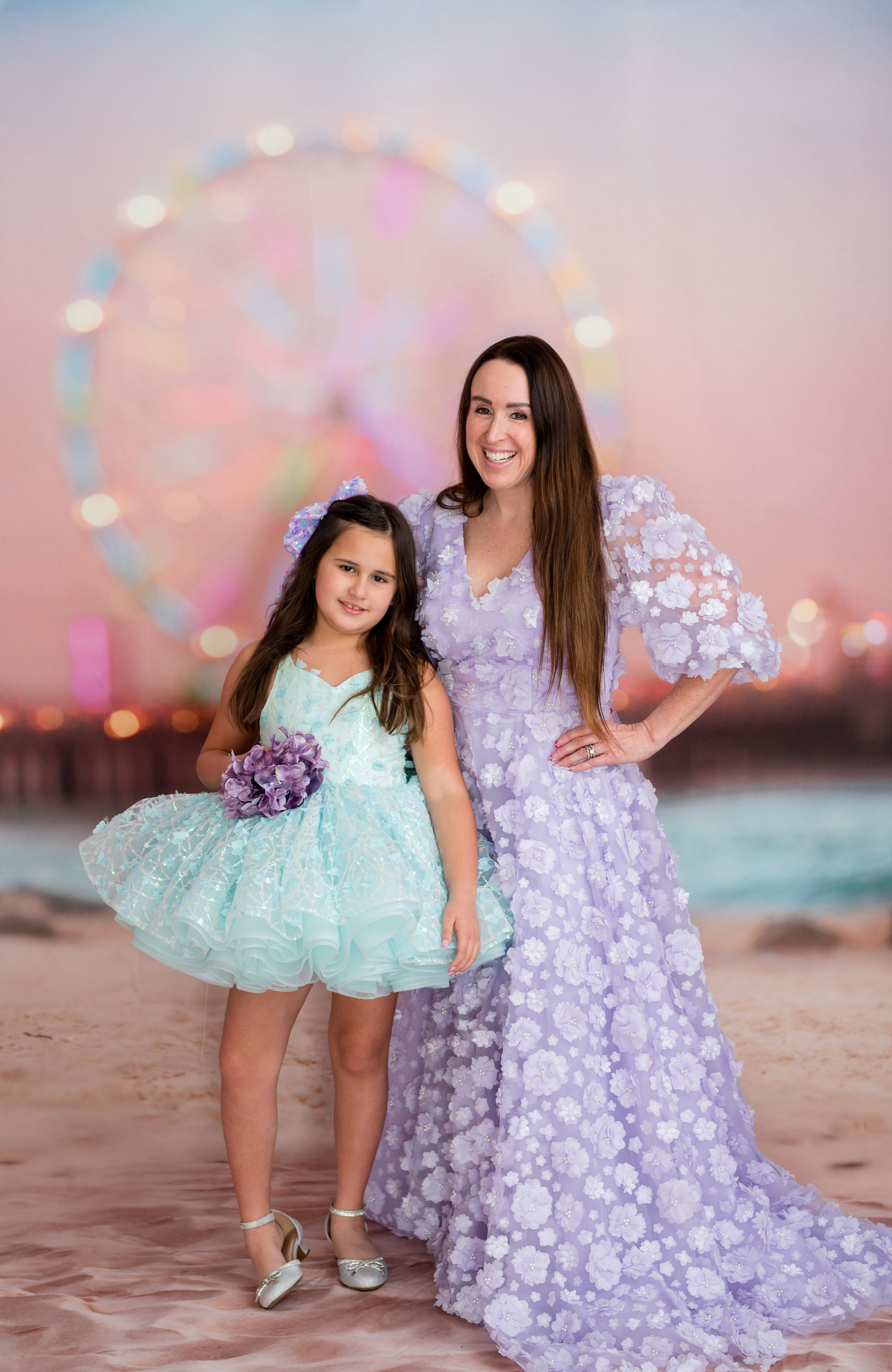 Woman and young girl in floral dresses standing on a beach with a Ferris wheel in the background.