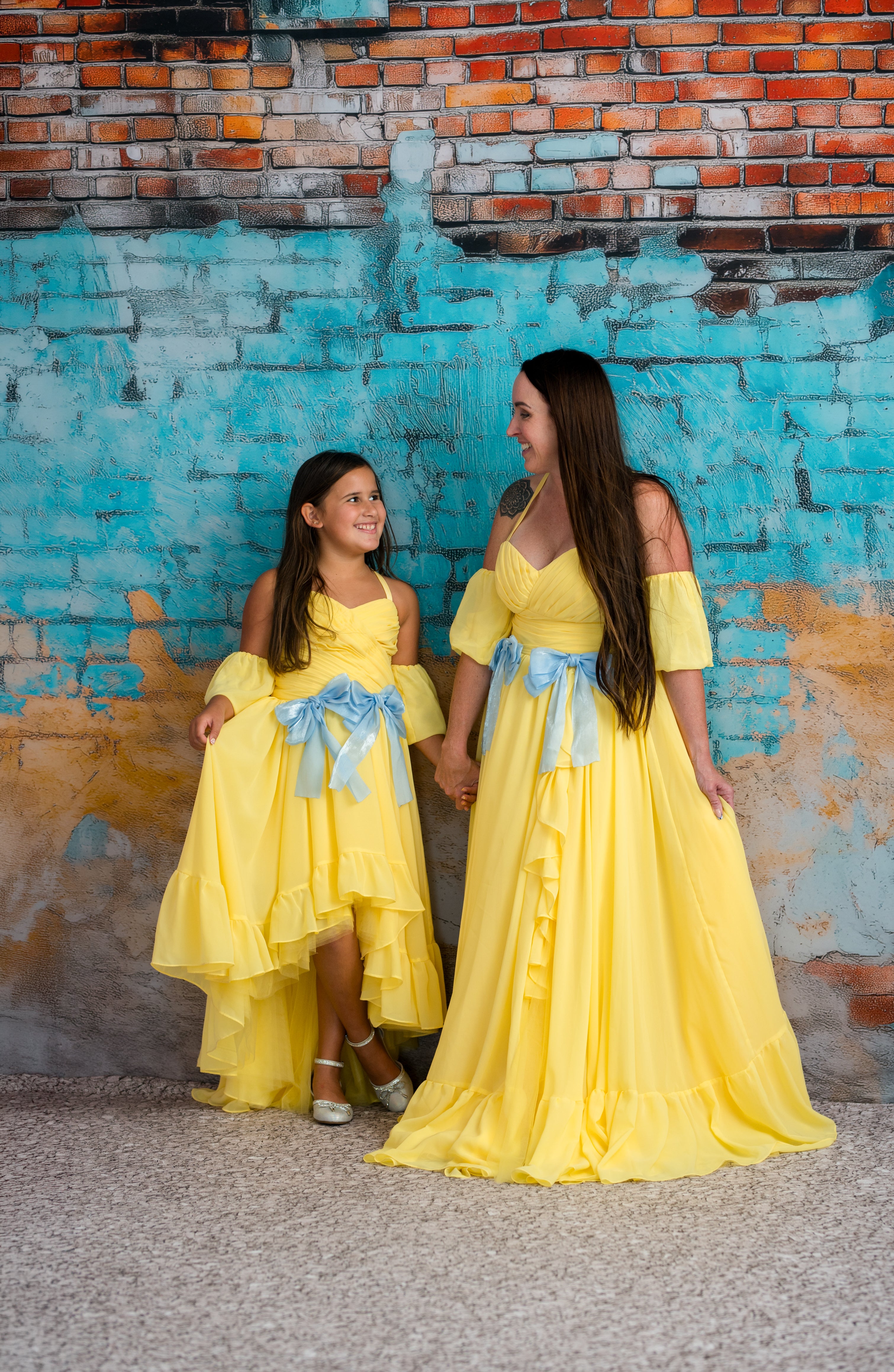 Two women in yellow dresses standing against a textured wall.