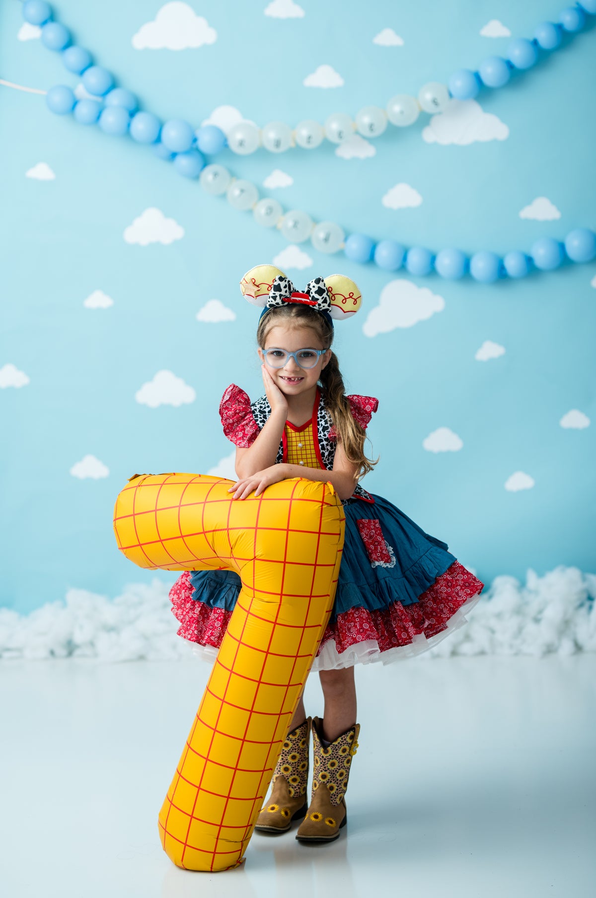 A young girl in a Woody-inspired costume stands behind a large yellow inflatable toy, posing in front of a light blue backdrop decorated with white clouds and blue balloon garlands.