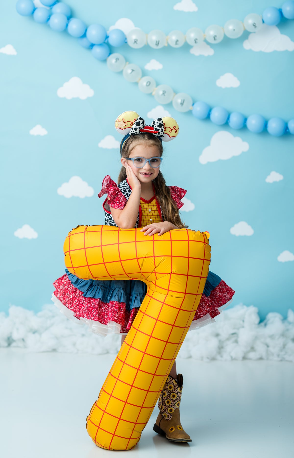 A young girl in a Woody-inspired costume sits on a large yellow inflatable block against a blue sky-themed backdrop with white clouds and balloon garlands.