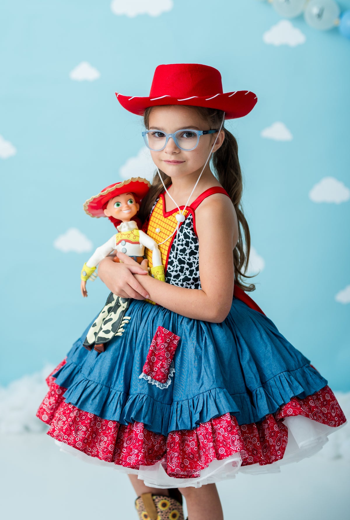 A young girl wearing a Woody-inspired denim cowboy dress with red patterned ruffles, a red hat, and glasses, holding a doll against a light blue cloud-patterned backdrop.